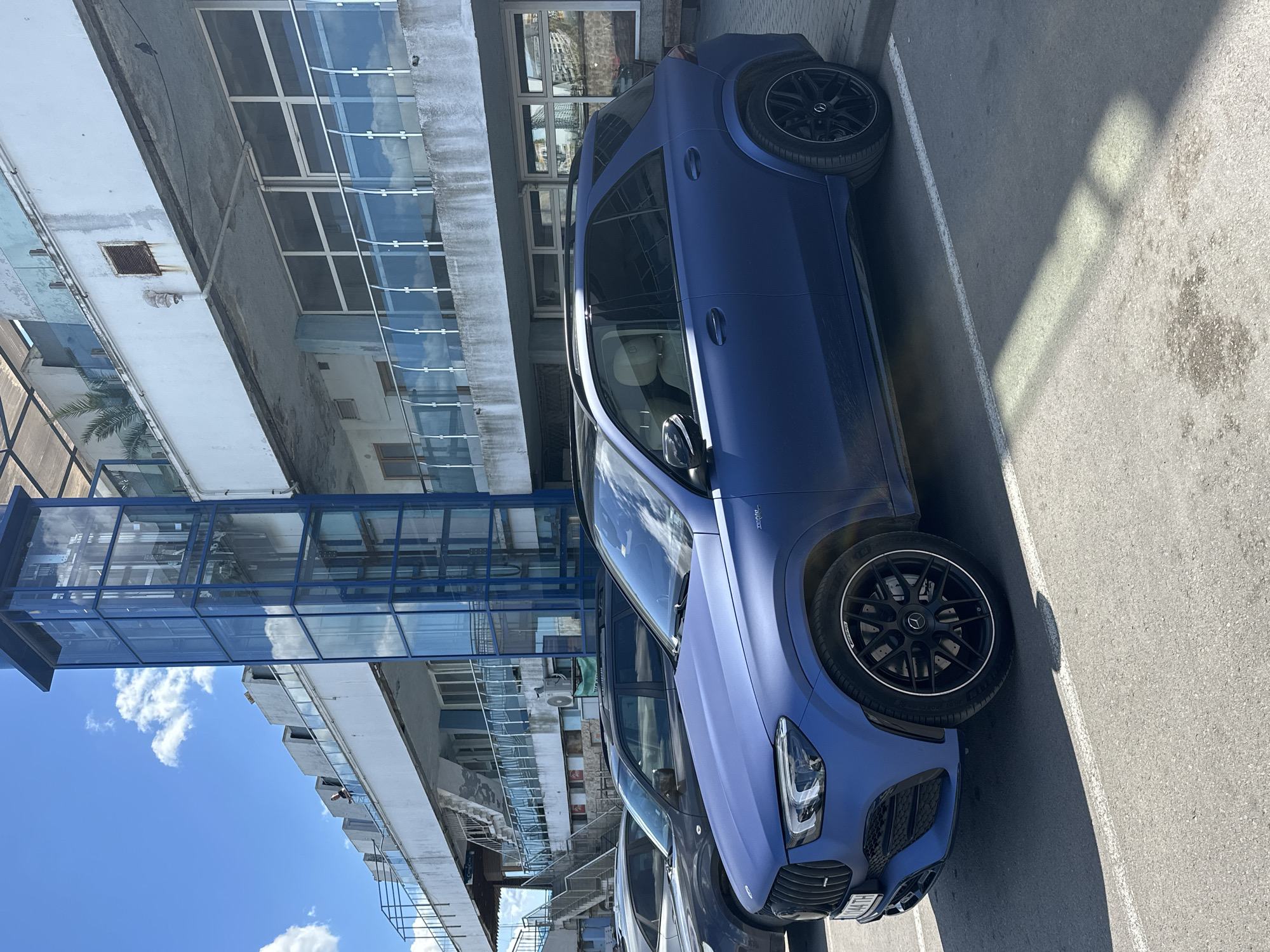 The image shows a blue Mercedes-Benz GLE AMG parked in an outdoor parking area next to a building. The building has a modern architectural design with glass panels and walkways. The sky is visible with some clouds. The car appears to be well-maintained. The parking area is paved. The car is parked in Varna, Bulgaria.