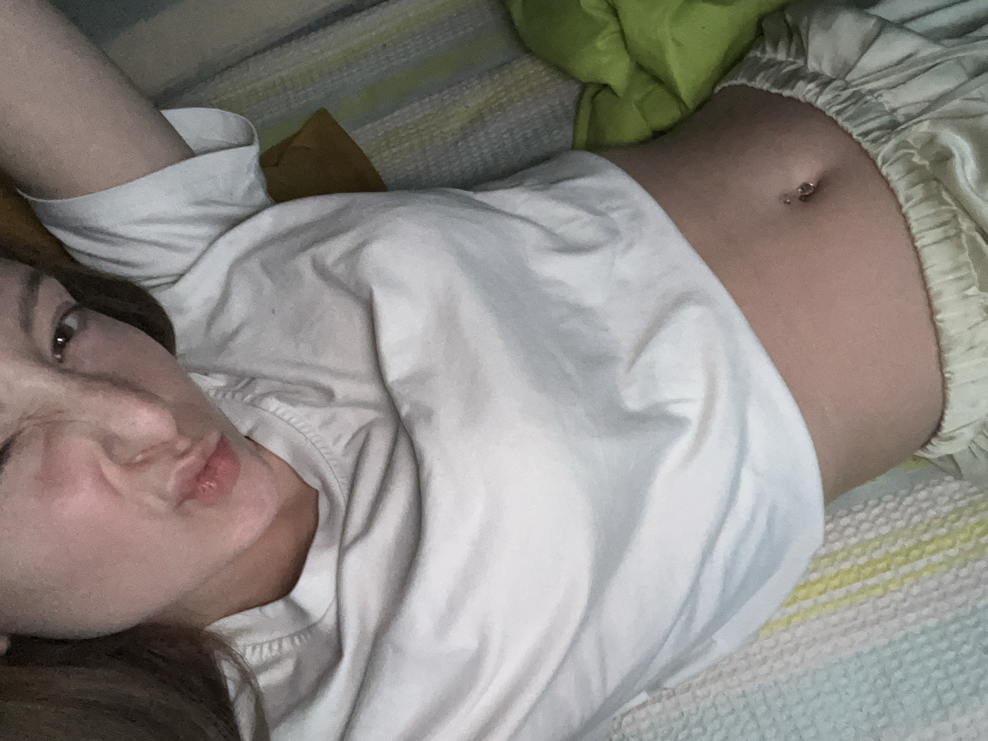 A woman is lying down, wearing a white shirt and light-colored pajama pants, with a visible navel piercing. She is making a kissing face towards the camera. The background shows a striped mattress and a green pillow. The location is not evident from the image and cannot be determined to be Varna, Bulgaria.