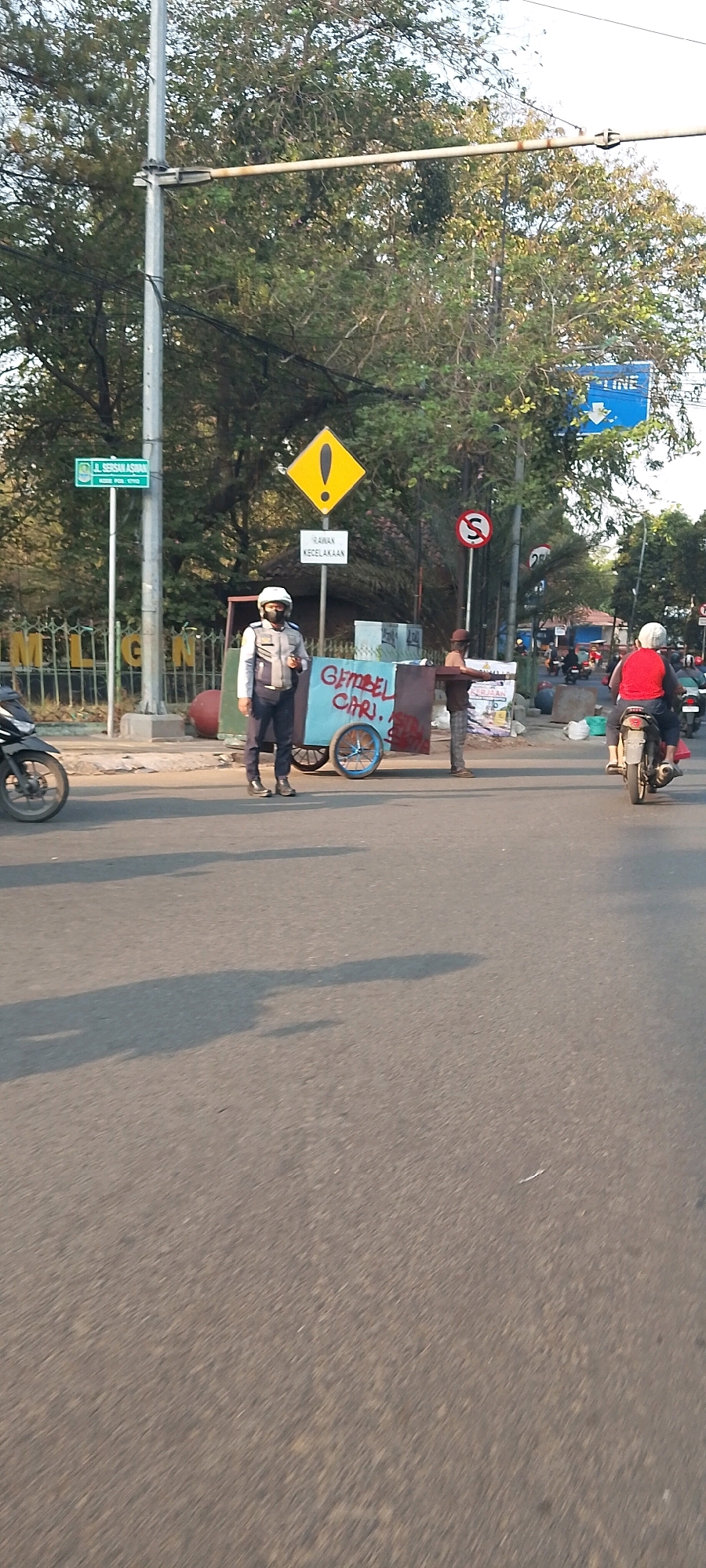 A traffic policeman wearing a white helmet, face mask, and a light-colored uniform stands on the asphalt road in front of a pushcart with a light blue body and red trim. The cart has a handwritten sign on its side that reads "GEMBEL CARI?" The policeman's right arm is extended, possibly directing traffic. Behind him, a person in a dark shirt and pants stands with their arms extended sideways, also potentially involved in traffic management. The pushcart has two blue wheels. In the background, trees with lush green foliage line the street. Several traffic signs are visible: a yellow diamond-shaped sign with an exclamation mark, a sign with the text "RAWAN KECELAKAAN" (accident-prone), and a circular sign with a red border and a black "S" inside. A blue sign with white text and an arrow pointing down reads "GASOLINE LINE". Further in the background, other people and motorcycles are present, indicating a moderately busy street scene in a city. The ground is paved with asphalt. The sunlight casts long shadows across the road, suggesting it's late afternoon. Based on the presence of the "GASOLINE LINE" sign and the general urban environment, this scene is likely located in Bekasi, Indonesia.