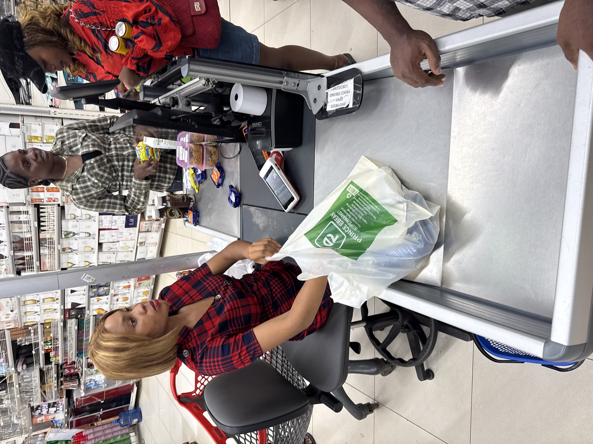 The image shows the interior of Prince Ebeano Supermarket, a retail establishment located in Lokogoma, Abuja, Nigeria. A woman with blonde hair and a red plaid shirt sits at a checkout counter, packing groceries into a plastic bag bearing the supermarket's name. Two other individuals are present: one in a red patterned top and the other in a green plaid shirt, both appearing to be customers at the checkout. A male is present. The checkout area includes a point-of-sale system with a monitor, scanner, and receipt printer. Shelves stocked with a variety of products are visible in the background.