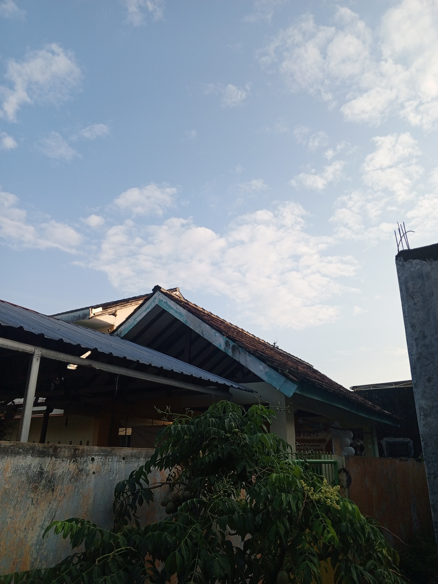 The image captures a scene in Sumbersari, Indonesia, with a clear blue sky dotted with scattered white clouds. In the foreground, a partially overgrown wall shows signs of wear and tear. Adjacent to the wall, a leafy tree with small green fruits or flowers branches into the frame. Behind the wall, a building with a tiled roof is visible, featuring an overhanging corrugated metal roof extension. Another building structure, a tall concrete pillar with exposed rebar, stands on the right side of the image. The overall impression is one of a residential or community area under a bright sky, with elements of both established structures and natural growth.