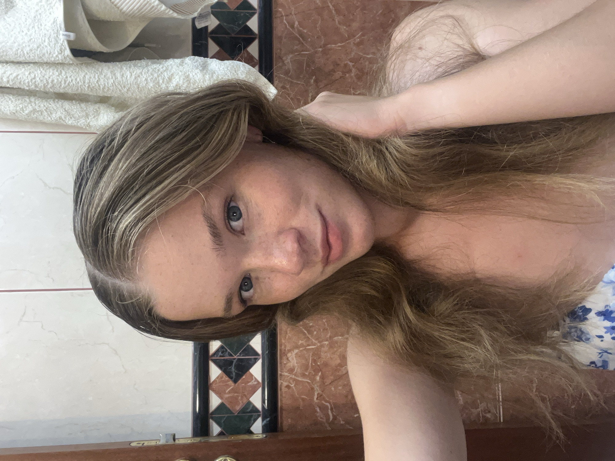 A young woman with long, wavy, blonde hair and blue eyes, wearing a white and blue floral top, poses for a selfie in a bathroom. The woman has freckles and is holding her hair with her right hand. The background shows a tiled wall with a marble pattern and a white towel hanging. The scene is a bathroom in Budva, Montenegro.