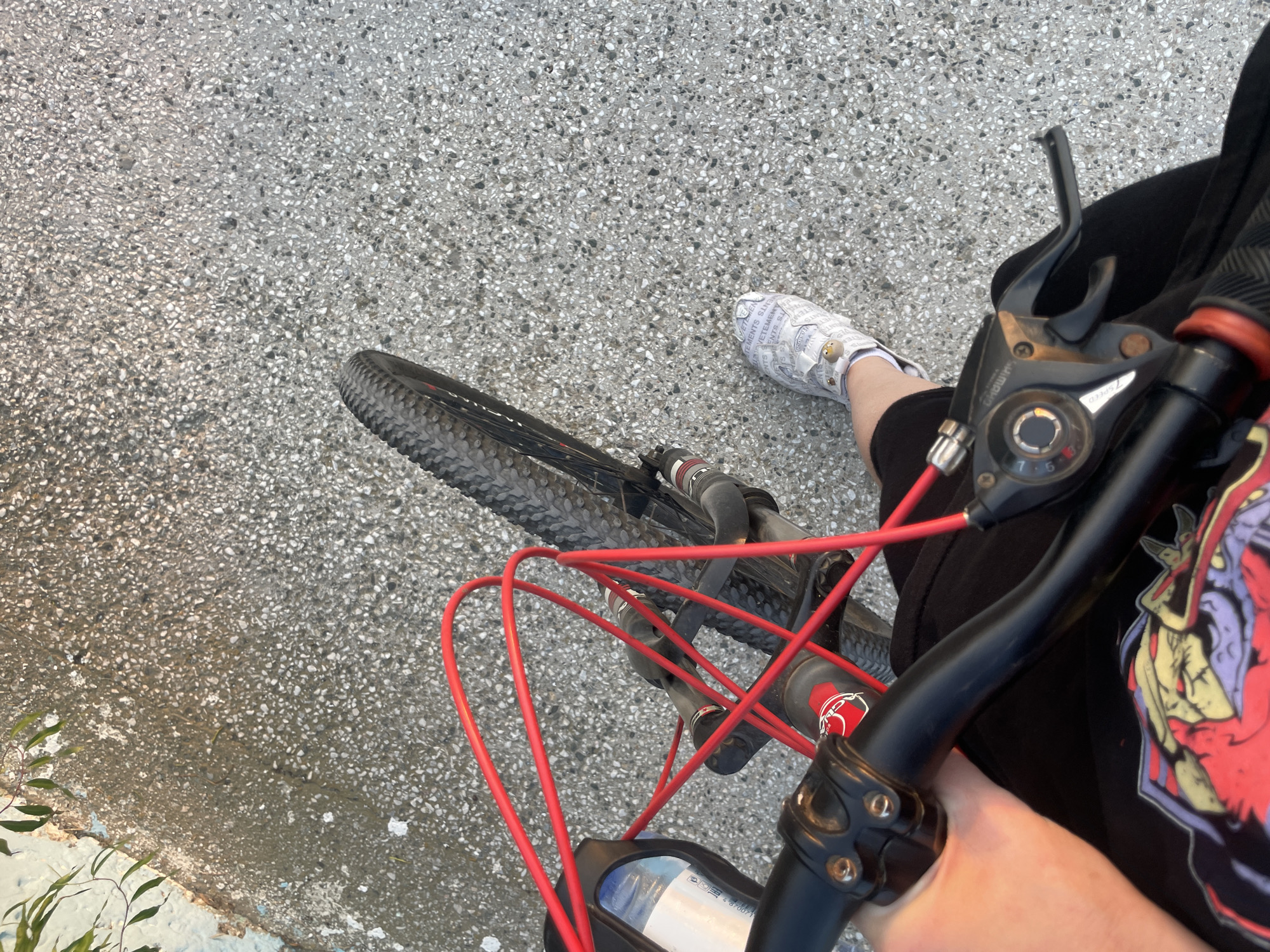 The image is a first-person perspective shot taken from a bicycle, likely in Aktau, Kazakhstan. The foreground shows a person's left hand gripping the handlebars of the bicycle. The handlebars are black with red cable housing. A black t-shirt with a red and yellow graphic is visible on the person's lap. To the right, a white shoe with a repeating pattern of text and small cartoon characters is worn on the person's right foot. The shoe is patterned with the word "VETEMENTS". The bicycle's front wheel, with a knobby tire, is positioned on the right side of the frame, partially obscuring the ground. The ground itself is paved with a rough, speckled aggregate material. In the upper left corner, a blurry branch with green leaves hangs over a textured, light-colored surface, possibly a wall or curb. The lighting suggests it is daytime, with some shadows indicating the sun is at an angle. The scene depicts a person on a bicycle, possibly in an urban or suburban outdoor setting.