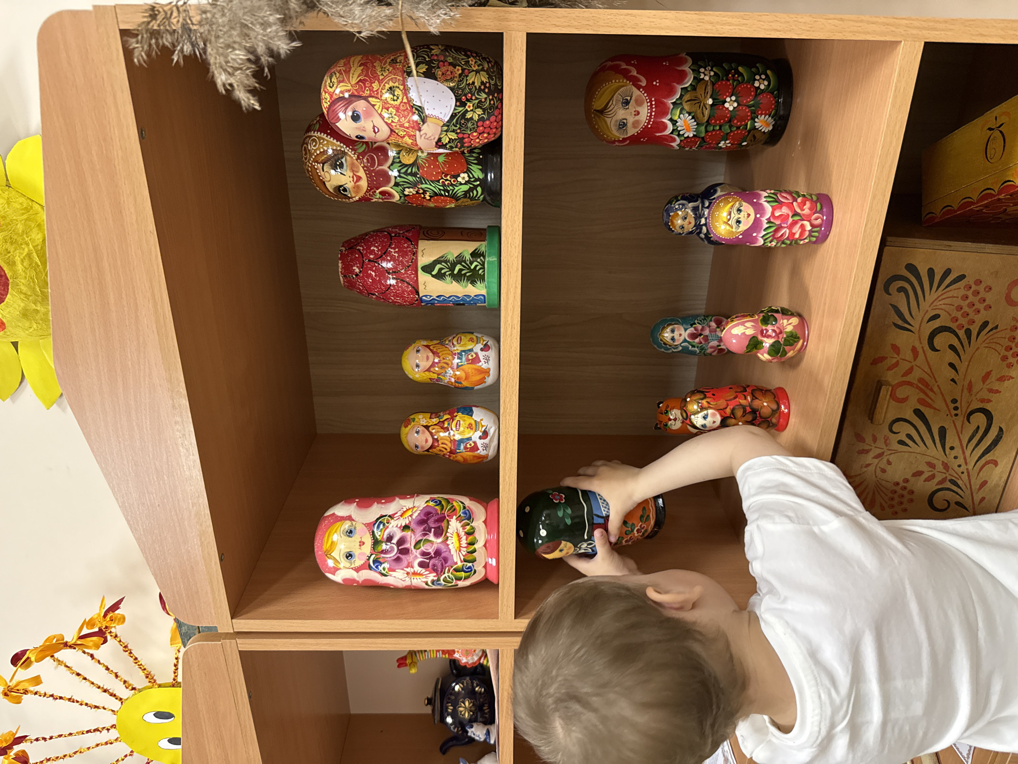 A young child in a white shirt is interacting with a shelf filled with Russian nesting dolls (Matryoshka dolls). The child is reaching into the shelf, seemingly placing or arranging the dolls. The shelf is made of light-colored wood and has multiple compartments. The dolls are of varying sizes and are decorated with traditional Russian patterns and motifs. A decorated wooden box is visible to the side of the shelf. The exact location is not discernible from the image, other than based on the question, Шебунино, Russia.
