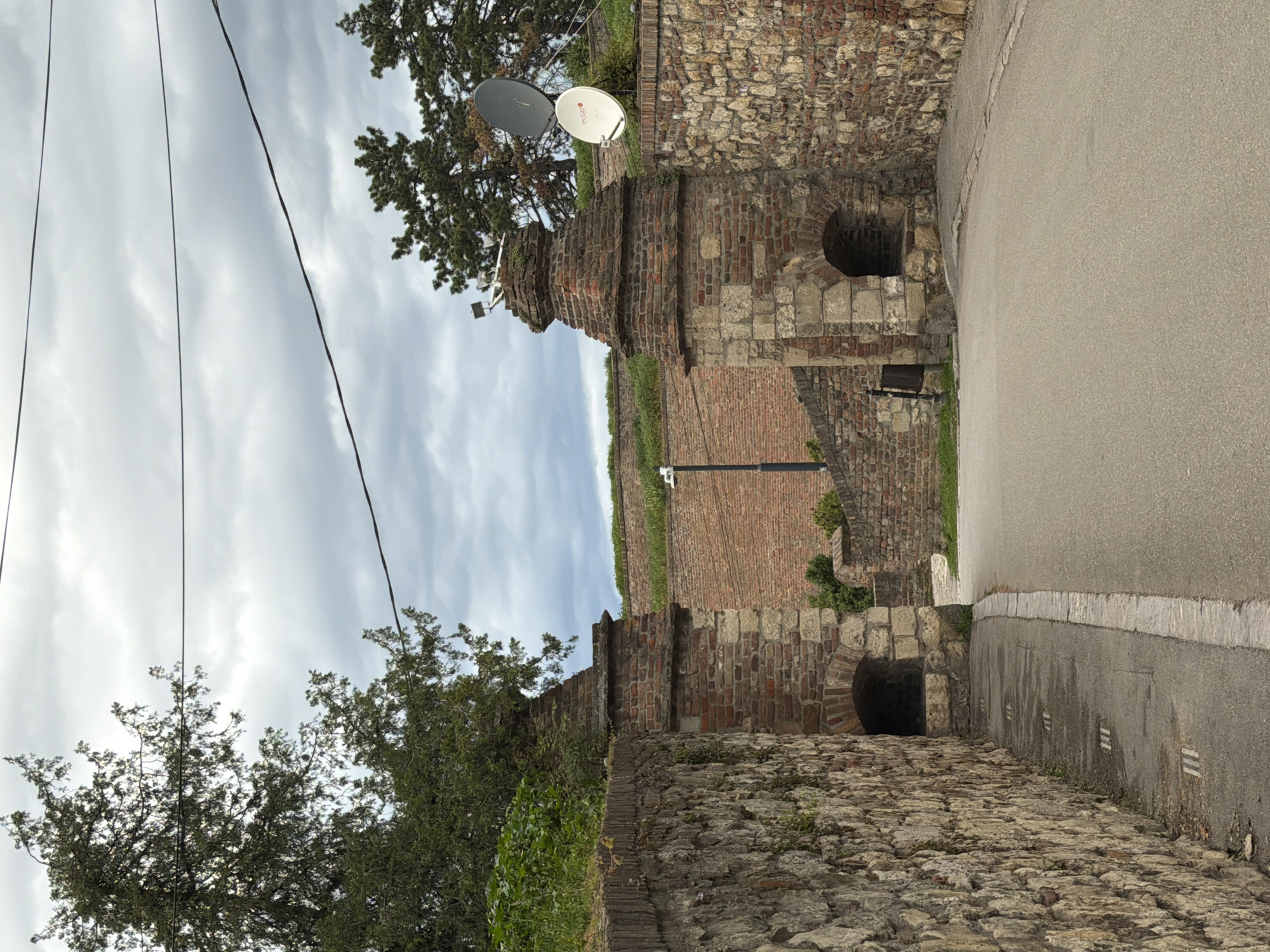 The image captures an outdoor scene within a historical setting, likely the Belgrade Fortress in Serbia, characterized by ancient fortifications under an overcast sky.
The scene features a narrow, paved road or pathway winding between old, rugged stone and brick walls. On the left side of the frame, a substantial wall composed of varied stone blocks and reddish-brown bricks rises, partially obscured by dense green trees and foliage. Several thin, dark utility wires stretch across the light gray, cloudy sky overhead. High on the left, two white and gray satellite dishes are mounted on a structure or tree, with the letters "TSA" clearly visible on one of them.
To the right, the asphalt-paved road curves gently, bordered by another, lower wall constructed from a mix of stone and brickwork. This wall contains at least two dark, arched openings, resembling tunnels or small alcoves. Further into the background on the right, another tall brick wall ascends, reinforcing the impression of a fortified structure. A slender, black pole, possibly a lamppost, stands near the center of the image.
The weather is overcast with a uniformly light gray, cloudy sky, suggesting a daytime scene without direct sunlight. There are no people visible, contributing to a quiet, timeless atmosphere. The architecture, including the aged stone and brickwork, arches, and walls, strongly indicates a historical fortress or ancient city defense system. No specific activity or event is depicted.