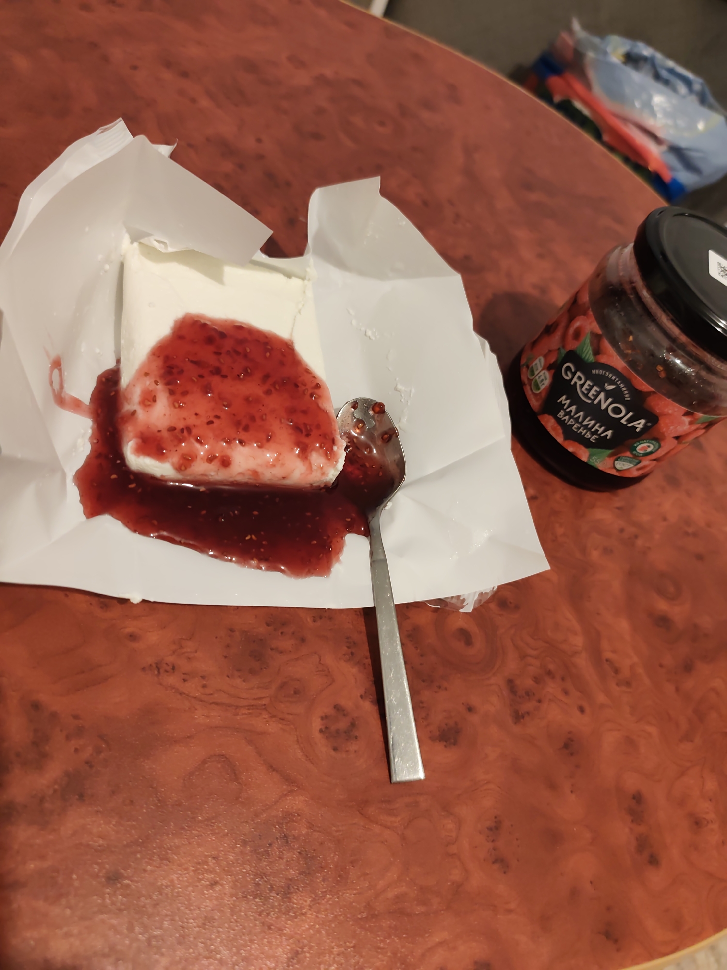 The image presents an indoor scene on a polished reddish-brown table, likely within a private residence in Chaikovsky, Russia. The central subject is a rectangular block of white, soft food, possibly curd cheese or a similar dairy product, resting on its partially unwrapped white paper packaging. A generous quantity of vibrant red raspberry jam, containing visible seeds and fruit pieces, is spread over the top surface of the white block and has spilled onto the surrounding paper and the table. To the right of the food item, a silver-colored metal spoon lies partially submerged in the spilled jam. In the upper right portion of the image, a clear glass jar with a black lid is visible. The jar's label displays "GREENOLA" and "МАЛИНА ВАРЕНЬЕ" (Malina Varenye, or Raspberry Jam in Russian), along with images of raspberries. The background, out of focus, shows indistinct colorful objects, possibly bags or other household items. No individuals are present in the frame.