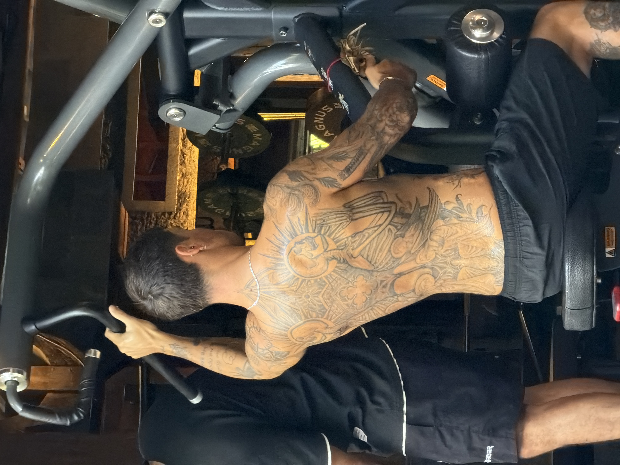 A shirtless man with extensive tattoos on his torso, arms, and back is exercising on a seated leg extension machine. He is wearing black shorts. The machine is grey with black padding. The setting is possibly a gym in North Kuta, Indonesia, based on the presence of "Magnus" weight plates, a brand associated with that location.
