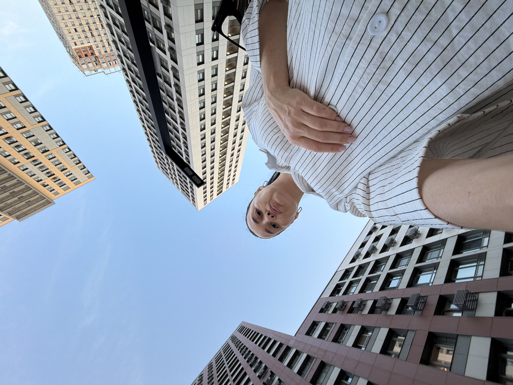 A low-angle, wide-shot selfie is captured in an urban environment with a clear blue sky. The composition is dominated by the sky and the facades of several modern, multi-story buildings that appear to be residential or office complexes. The buildings are made of various materials, including reddish-brown panels, grey concrete, and white cladding, with many windows visible.
In the foreground, a woman is positioned on the right side of the frame, looking upwards towards the camera. She has dark hair pulled back and is wearing a white shirt with thin black vertical pinstripes. Her left hand rests on her chest. The camera appears to be held at a very low angle, pointing upwards towards the woman and the buildings.
The image appears to have been taken in Moscow, Russia, likely in a modern residential district. The architectural style of the buildings, with their repetitive window patterns and varied cladding, is characteristic of contemporary urban development in Russian cities. No specific landmarks are identifiable that would pinpoint the exact location within Moscow, but the overall impression is of a built-up, modern city scape.