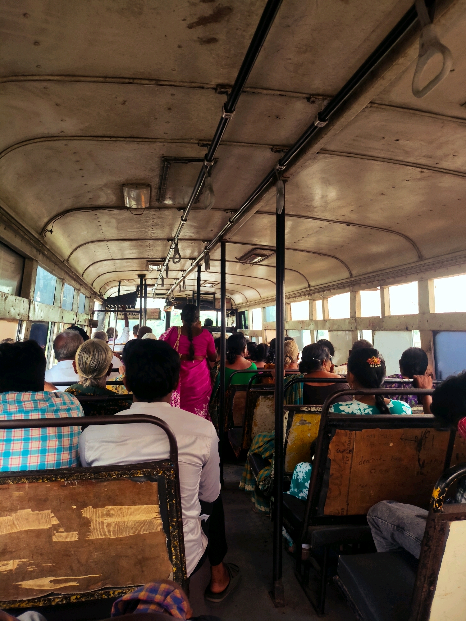 The image shows the interior of a bus filled with passengers, viewed from the rear. The bus appears old and worn, with a metallic ceiling and visible support poles. Passengers are seated on both sides of the aisle, facing forward. Several individuals are visible, including a woman in a bright pink sari standing in the aisle, appearing to be a conductor or passenger moving through the bus. Others are seated, some looking forward, others slightly to the side. The windows lining the bus are large and some show bright sunlight, suggesting it is daytime. The seats appear to be made of a dark material with worn wooden or cardboard backing. Some seats have metal frames. The floor of the bus is not clearly visible. The overall impression is of a crowded public transport vehicle in a warm climate. The location is identified as Budampadu, India.