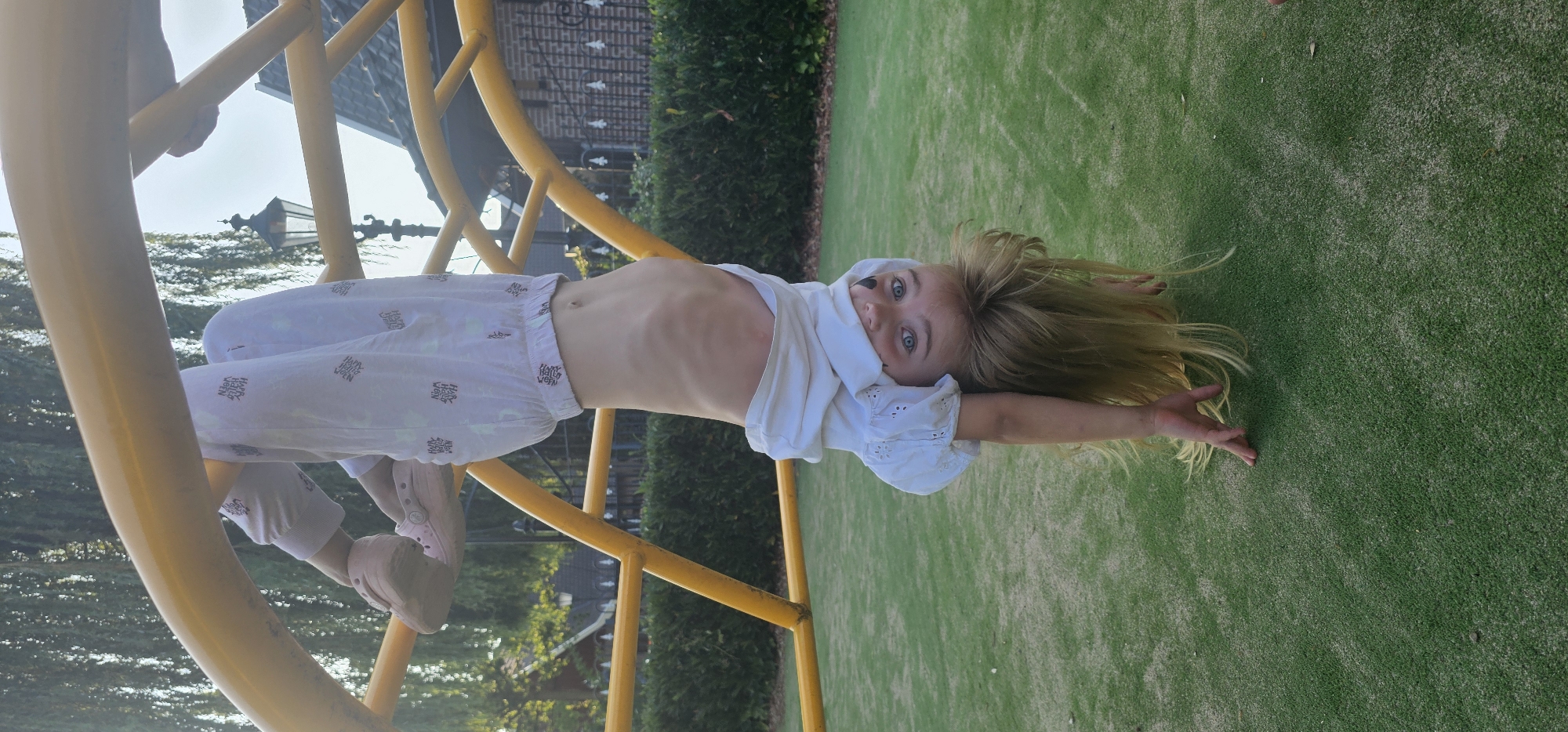 A young girl with blonde hair and blue eyes is upside down on a yellow jungle gym in a park, likely in Groenlo, Netherlands. She is wearing a white t-shirt and white pants with a pattern. Her arms are outstretched, and her hands are touching the green, artificial turf ground. The background shows trees, a street lamp, and a building. The lighting suggests it is daytime, with the sun casting shadows.