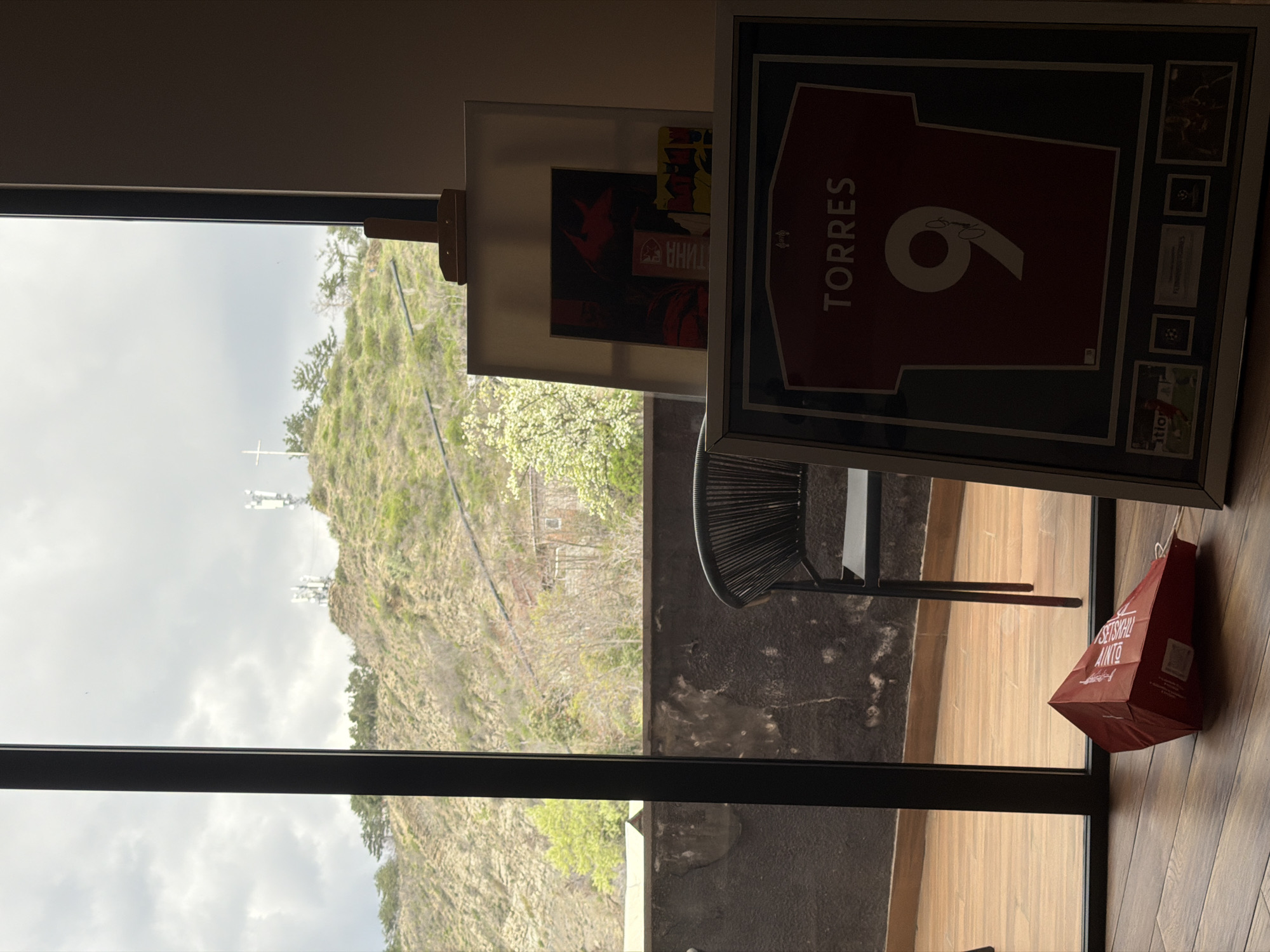 The image shows an interior scene with a view through a window to a hill, likely in the city of Tbilisi, Georgia. A framed football jersey with the name "TORRES" and the number "9" is leaning against the wall. To the left of the jersey is an artwork propped on a small easel. A dark wicker chair is placed between the artwork and the jersey. A red shopping bag with white text is also leaning against the wall next to the jersey. The environment outside the window shows a cloudy sky and a hill with trees, power lines and a white cross at the top.
