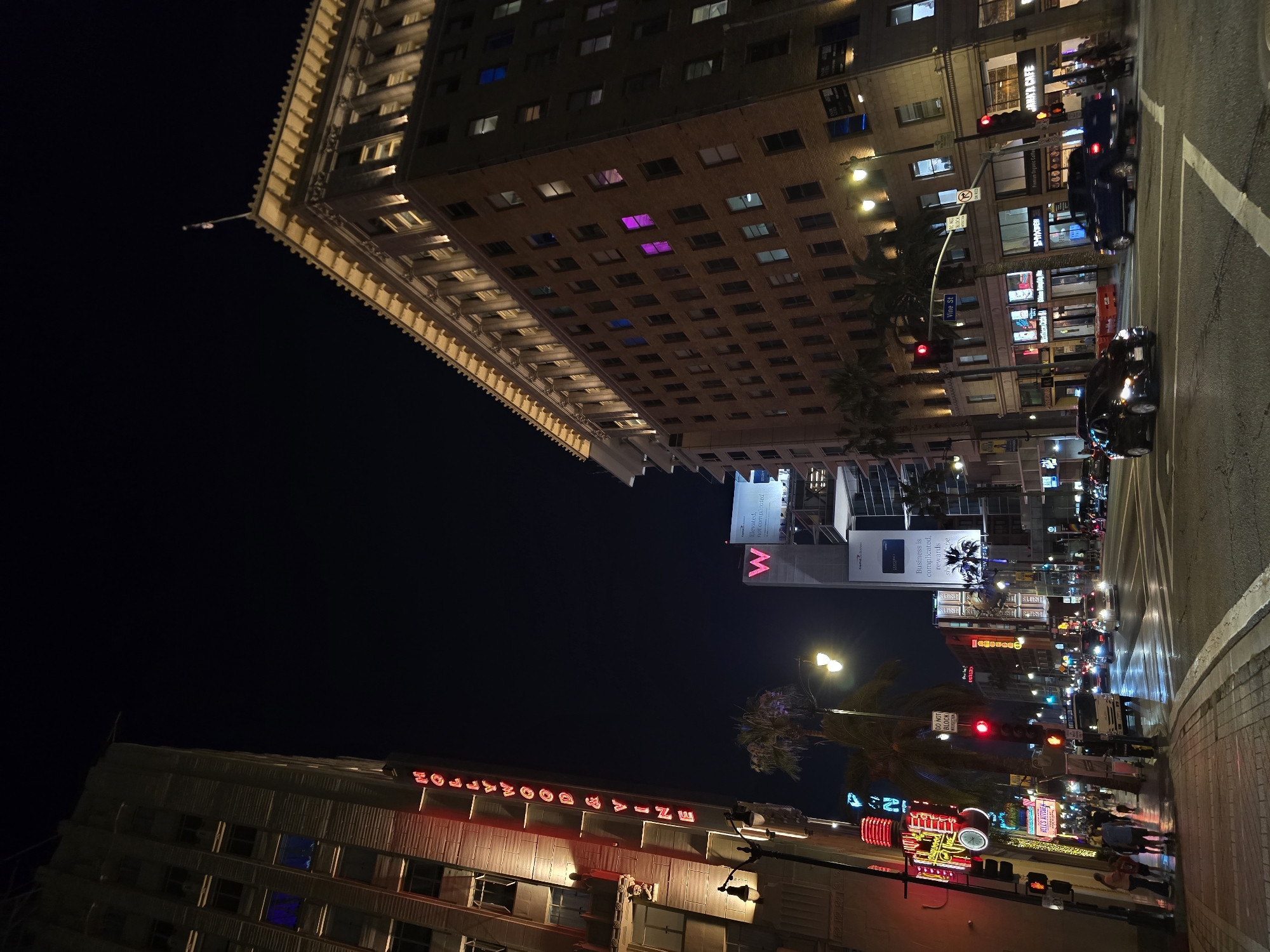 This is a night-time, medium-angle, full shot of the intersection of Hollywood Boulevard and Vine Street in Los Angeles, California. The image captures a lively urban scene with glowing neon signs, illuminated buildings, and a street intersection with traffic.
On the left side of the frame, a tall, light-colored building displays a vertical neon sign that reads "HOLLYWOOD & VINE" in red lettering. Another neon sign below it is partially visible and features the words "HOLLYWOOD BLVD". Streetlights and traffic lights add to the illumination of the scene, with red lights prominently displayed on the traffic signals.
The right side of the image is dominated by a large, multi-story brick building with rows of lit windows. Some windows are illuminated with colorful lights, including pink and blue. Another modern, white building with a large "W" logo is visible behind it.
The street is wet, reflecting the lights, suggesting recent rain. Several cars are driving through the intersection, and pedestrians are walking on the sidewalks, some of them silhouetted against the bright lights. Palm trees are scattered throughout the scene, adding a tropical element to the urban landscape. The overall atmosphere is vibrant and energetic, typical of a busy city intersection at night.