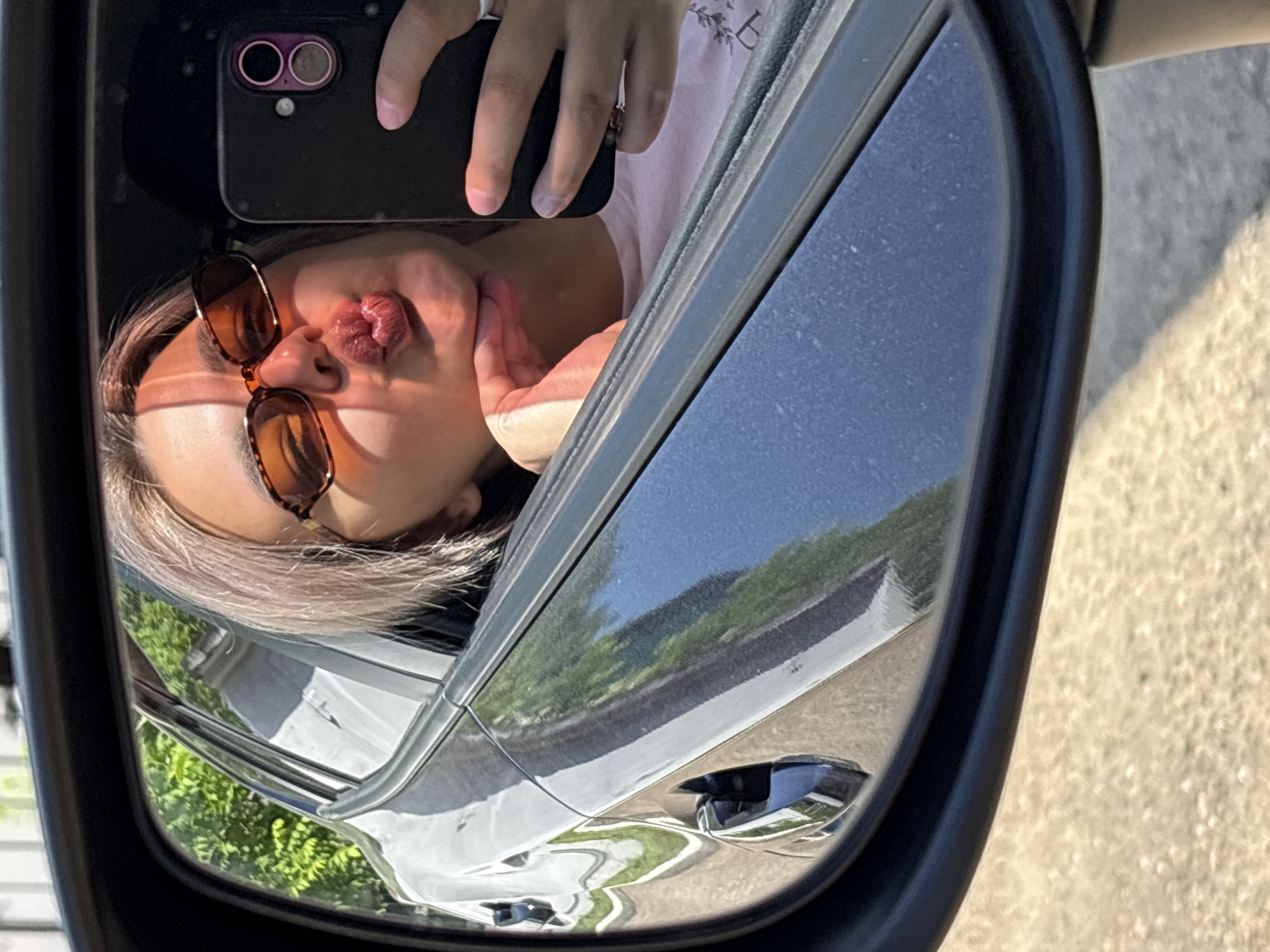 A woman with blonde hair and sunglasses is reflected in a car's side mirror, puckering her lips and holding a smartphone. The reflection shows greenery and a street. The image is taken from the perspective of the car's interior, looking out through the driver's side window and into the side mirror. The woman is wearing a pink shirt. The environment appears to be outdoors, possibly in a city, though no specific landmarks of Birobidzhan, Russia are visible.