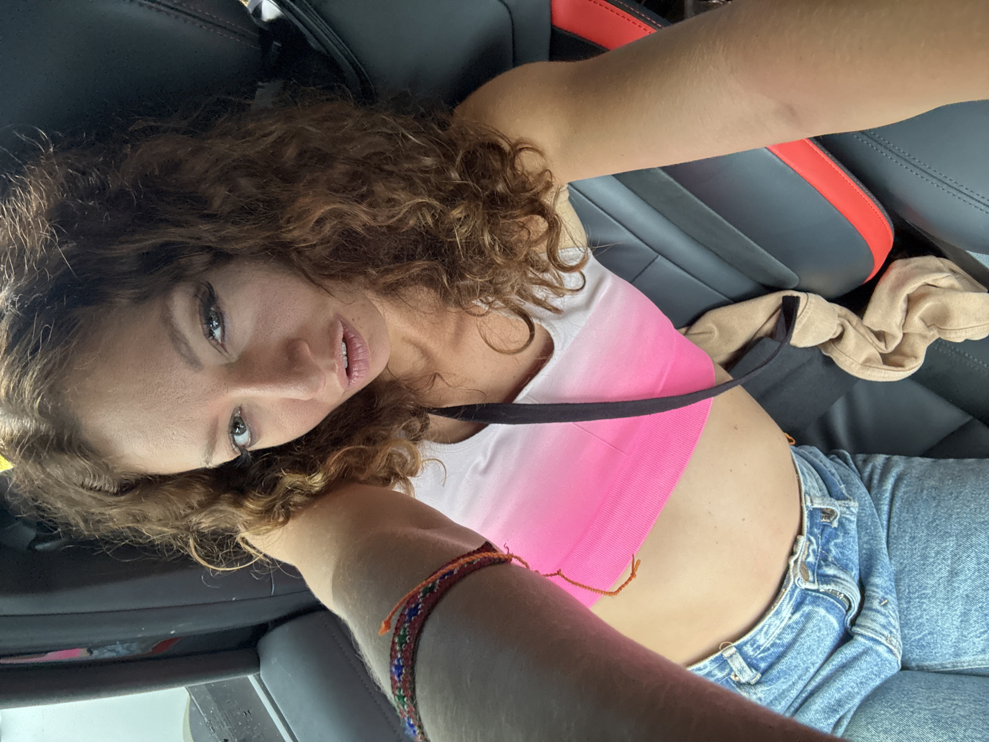 A woman with curly brown hair is seated in a car, wearing a pink and white sports bra and blue jeans. She is turned towards the camera and wearing a seatbelt. The interior of the car has black leather seats with a red accent on one seat. A tan piece of clothing is visible on the seat next to her. A colorful bracelet is on her arm. The location and details of Binh Lu, Vietnam, are not discernible from the image.