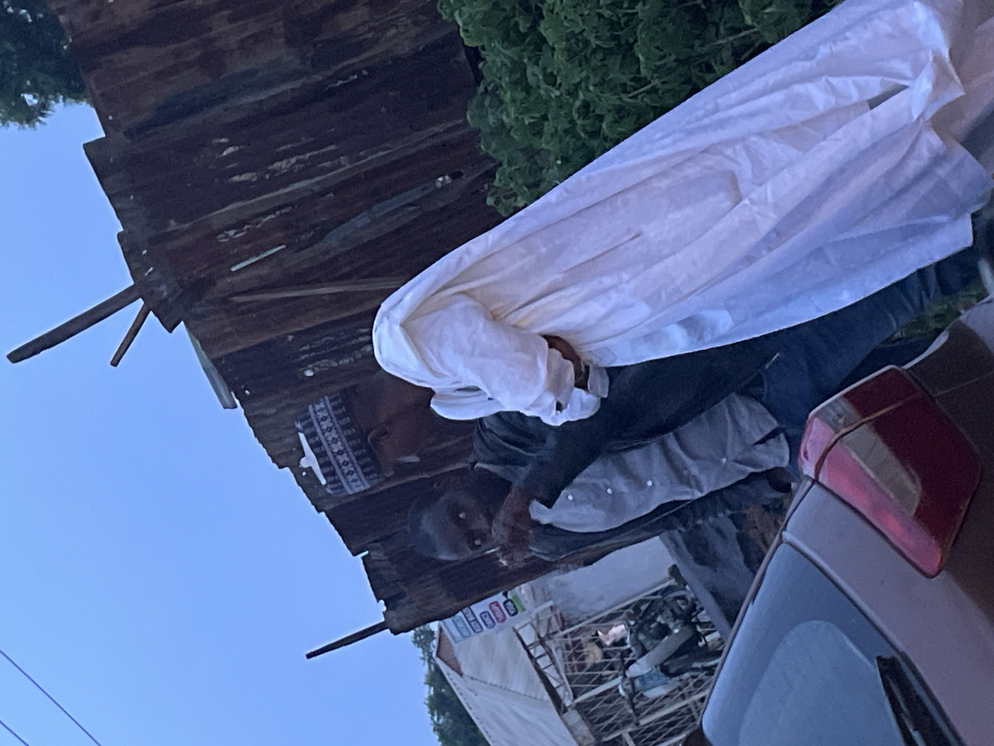 Two men are standing next to a car in an outdoor, urban environment. The man on the left is wearing a light-colored button-down shirt and a dark jacket. He has dark skin and hair and is looking towards the camera. The man on the right is wearing a traditional white robe and a patterned cap. He is facing away from the camera. In the background, there is a rusty metal fence, lush green foliage, a building with a striped awning, and a motorcycle parked near a grilled gate. A portion of a dark-colored car is visible in the foreground. The sky is overcast and the lighting suggests it is either early morning or late evening. The location appears to be the city of Jos, Nigeria.
