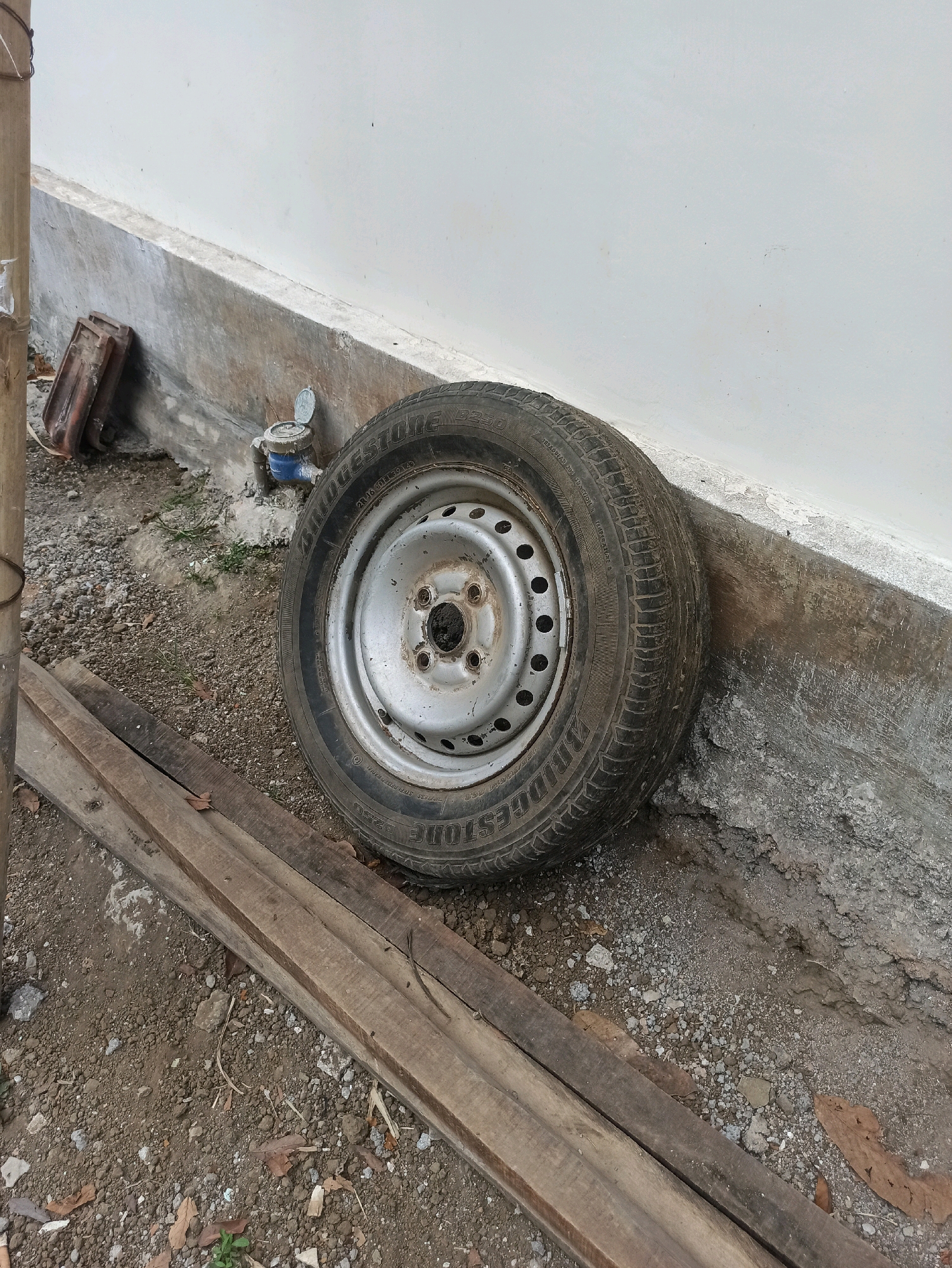 A single car tire rests against a white wall, leaning on a concrete foundation. The tire, with a silver rim, is positioned on a ground covered with gravel and scattered leaves. To the left of the tire, a section of a bamboo pole is visible. In the background, near the tire, a blue water meter with a silver lid is installed on the concrete. Two rectangular pieces of reddish-brown material, possibly tiles, are stacked near the wall, to the left of the water meter. In the foreground, two wooden planks lie horizontally on the gravelly ground. The scene appears to be an outdoor, unmaintained area, potentially a side yard or alleyway. The location is identified as Brambang Darusalam, Indonesia.