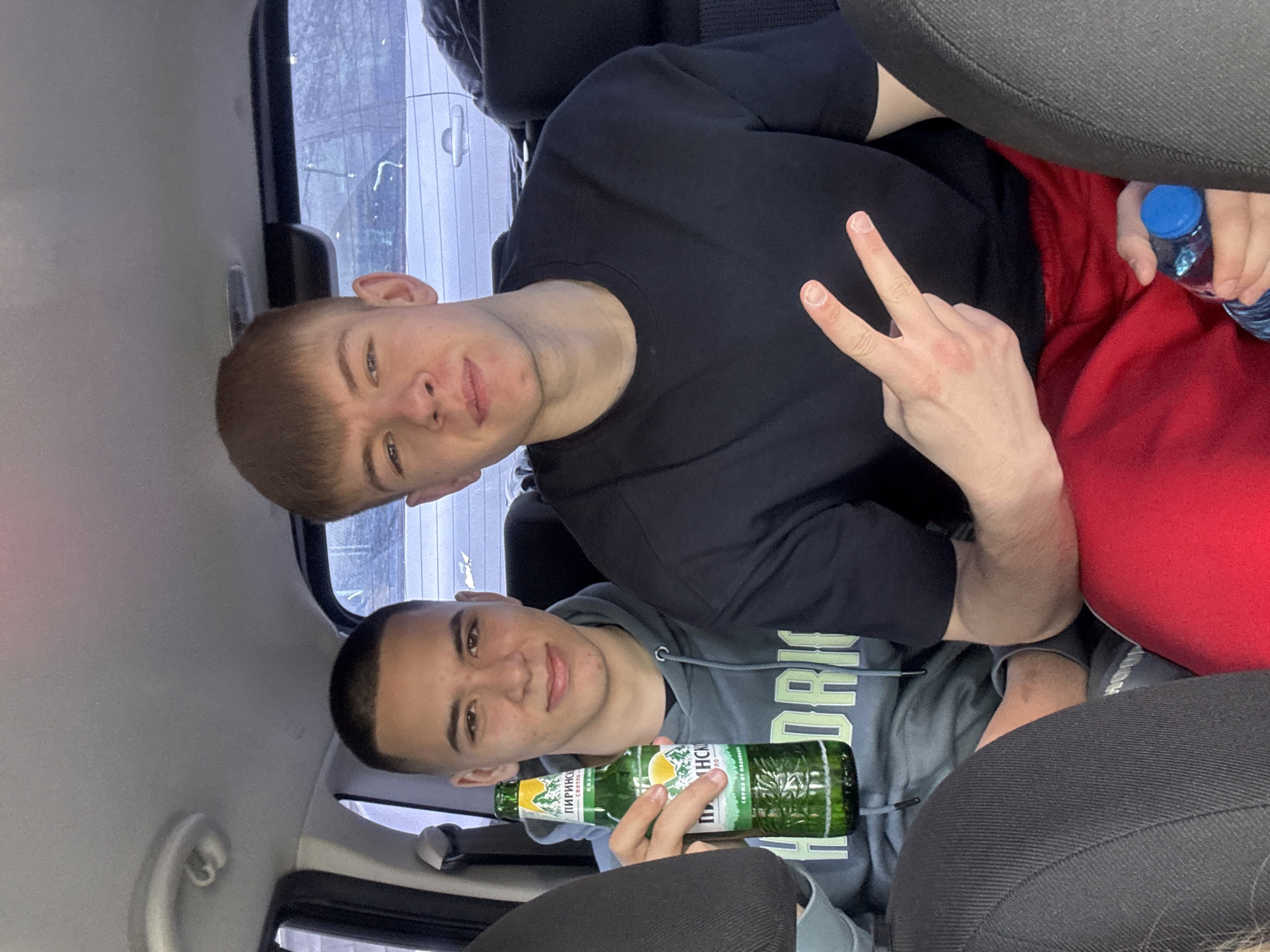 Two young men are sitting in the backseat of a car. The first man, positioned in the upper half of the frame, is wearing a black t-shirt and red sweatpants, and he is holding up a peace sign with his right hand. He also holds a bottle of water with a blue cap. The second man, in the lower half, is wearing a grey hoodie and holding a bottle of "Pirinsco Svetlo" beer. The setting appears to be the interior of a vehicle, with visible seats and windows. Based on the "Pirinsco Svetlo" beer bottle, the location is likely in Bulgaria, with Pirinsco brewery is located in Blagoevgrad, Bulgaria, close to Parvenets.