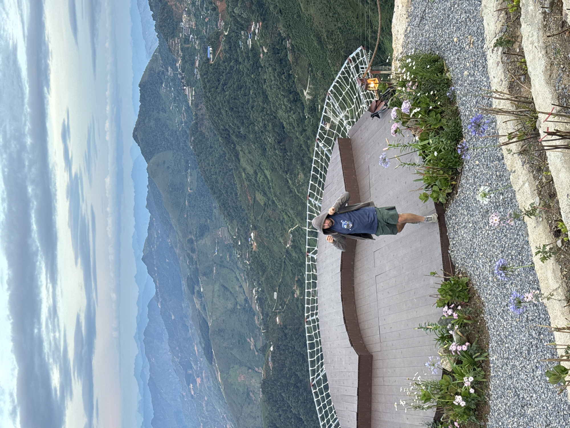 An outdoor, high-angle shot captures a male figure standing on a wooden viewing platform overlooking a vast mountainous landscape in Ta Xua, Vietnam. The man, positioned slightly left of center, wears a dark blue t-shirt with a light graphic, dark green shorts, light-colored athletic shoes, and a dark grey hooded jacket, which he appears to be adjusting with both hands near his shoulders. He has a beard and is looking slightly down and to his left.
The irregularly shaped platform, constructed from dark brown wooden planks, is equipped with a white safety net or rope barrier extending around its perimeter. A warm, illuminated lantern hangs near the right edge of the platform.
Beyond the platform, numerous layers of green, forested mountains extend into the hazy distance under an overcast sky with scattered grey clouds. The nearest mountainsides are densely covered in trees, while further peaks exhibit atmospheric perspective, appearing progressively lighter and bluer. Scattered houses and small settlements are visible on the mountain slopes, connected by winding dirt roads or paths. Some areas show patches of disturbed reddish soil, indicating cultivation or construction.
The immediate foreground features a grey gravel path surrounding the wooden platform. Within this gravel, and in tiered stone garden beds, various flowering plants are visible, including white, pink, and purple blooms, with prominent blue-purple flower clusters resembling Agapanthus. Some bare plant stalks are also present in the lowest foreground tier.