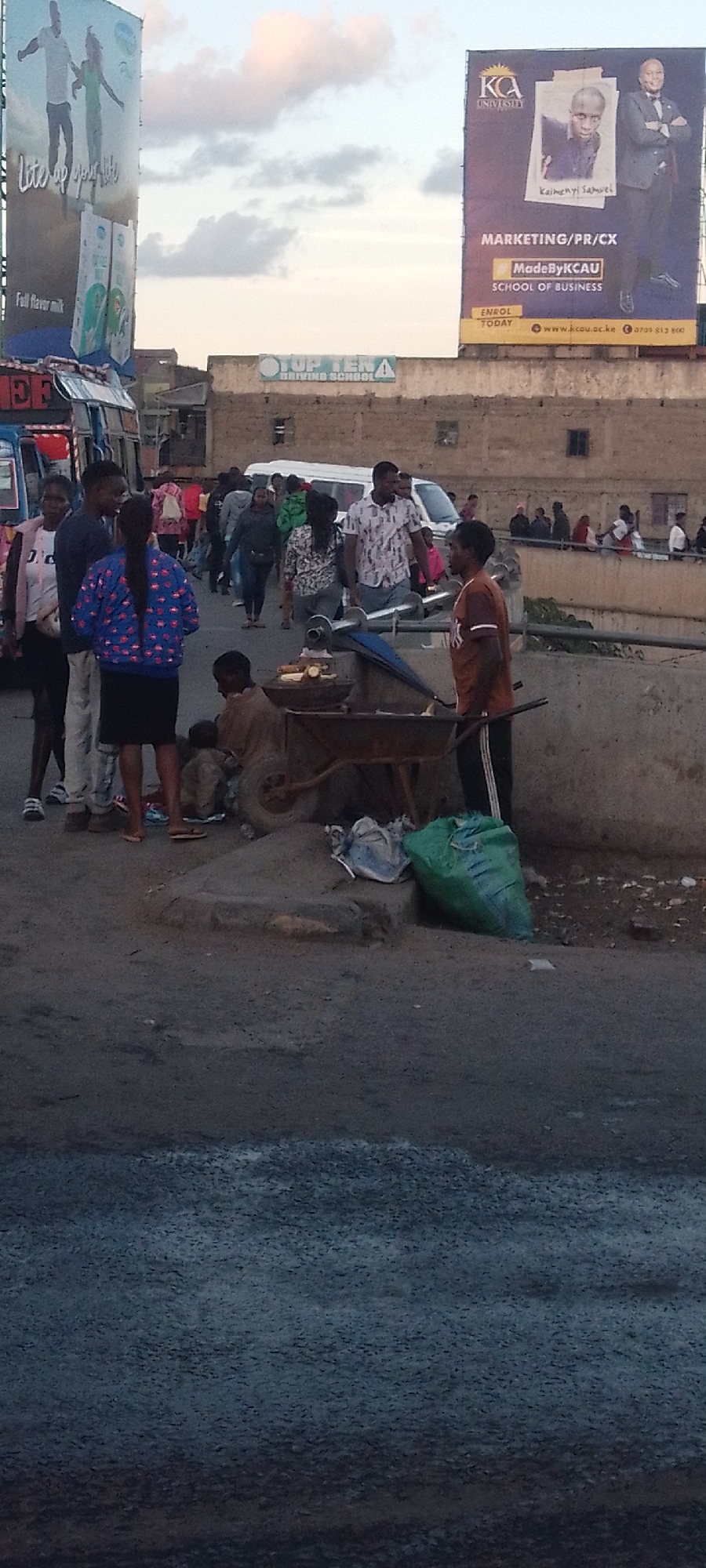 The image depicts a busy street scene in Nairobi, Kenya. Several people are gathered near a street vendor, who appears to be selling cooked corn. The vendor is standing next to a wheelbarrow, and a green bag is placed nearby. Some individuals are standing and talking, while others are walking along the street. In the background, there's a building with signs for "TOP TERA DRIVING SCHOOL" and "KCA University," with billboards advertising "Full Flavor Milk" and "Marketing/PR/CX MadeByKCAU School of Business." A white van is visible behind the crowd, and the overall environment suggests a bustling urban setting.