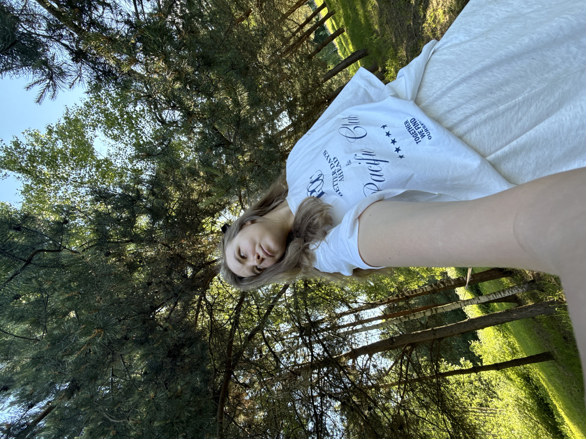 A low-angle, wide-lens selfie captures a young adult female, likely in her late teens or twenties, standing outdoors in a wooded environment. She possesses long, light brown hair, partially secured, and directs her gaze upwards and to her left (viewer's right) with a contemplative expression. Her right arm, holding the camera, extends into the lower foreground. She is dressed in a white, loose-fitting t-shirt prominently displaying blue text: "BETTER DAYS AHEAD" and "Pacific Club" in a script font, followed by five stars and "TOGETHER WE FIND OURSELVES". A light-colored skirt or dress is visible beneath the t-shirt.
The background consists of a dense arrangement of trees, including numerous coniferous pines with dark green needles and visible cones, intermingled with deciduous trees, some exhibiting characteristic white bark, likely birch. Patches of clear blue sky are discernible through the tree canopy. The ground is covered with green grass, illuminated by natural sunlight that creates dappled patterns and shadows across the foliage and terrain. The landscape behind the subject shows an upward slope with additional trees and lush green undergrowth. The scene is brightly lit by direct sunlight, indicating a clear day. This scene is located in Копнино, Russia.