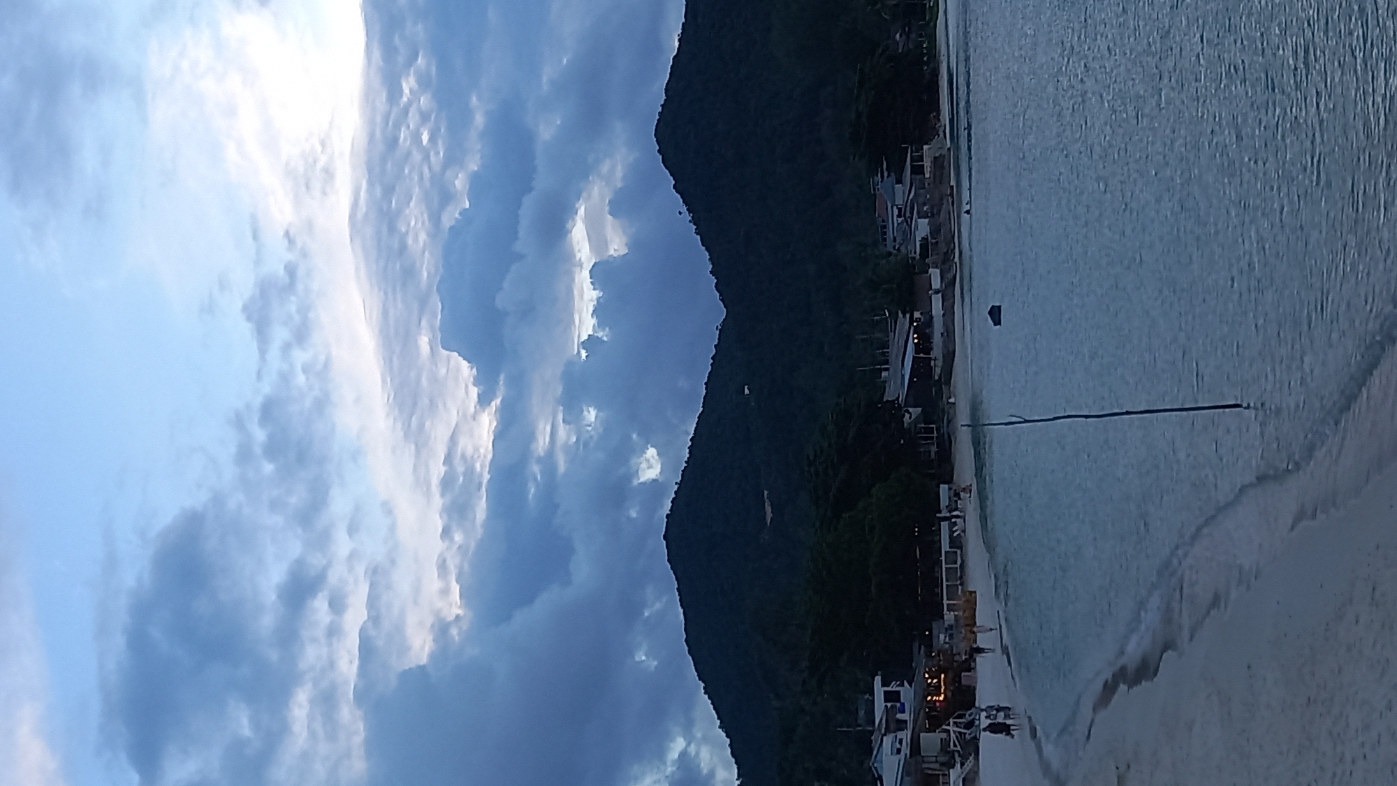 The image depicts a coastal scene on Ko Pha-ngan District, Thailand, likely during twilight or overcast conditions, as indicated by the muted light and dramatic cloud formations. In the foreground, a tranquil, light-colored sandy beach curves gently towards the viewer's left, bordered by the calm, pale blue sea. The water's surface is mostly smooth, with gentle ripples near the shore. A single, thin, dark stick or pole stands vertically in the shallow water, slightly off-center to the right.
Along the shoreline, a row of low-rise buildings, appearing to be shops or restaurants, are visible. Some have lights on, suggesting they are operational. Several small groups of people, appearing as indistinct figures, are walking on the beach and near the buildings.
In the background, a lush, dark green, mountainous landscape dominates the scene, rising steeply from the tree line behind the buildings. The mountains are covered in dense vegetation, and their peaks are silhouetted against the sky. The sky itself is a striking feature, filled with voluminous, heavy grey and blue clouds, with patches of lighter sky visible, hinting at breaks in the cloud cover or diffused sunlight. A small, dark, rectangular object, possibly a floating marker or a small boat, is visible in the water to the right of the central pole. The overall impression is one of natural beauty and coastal serenity under a dramatic sky.