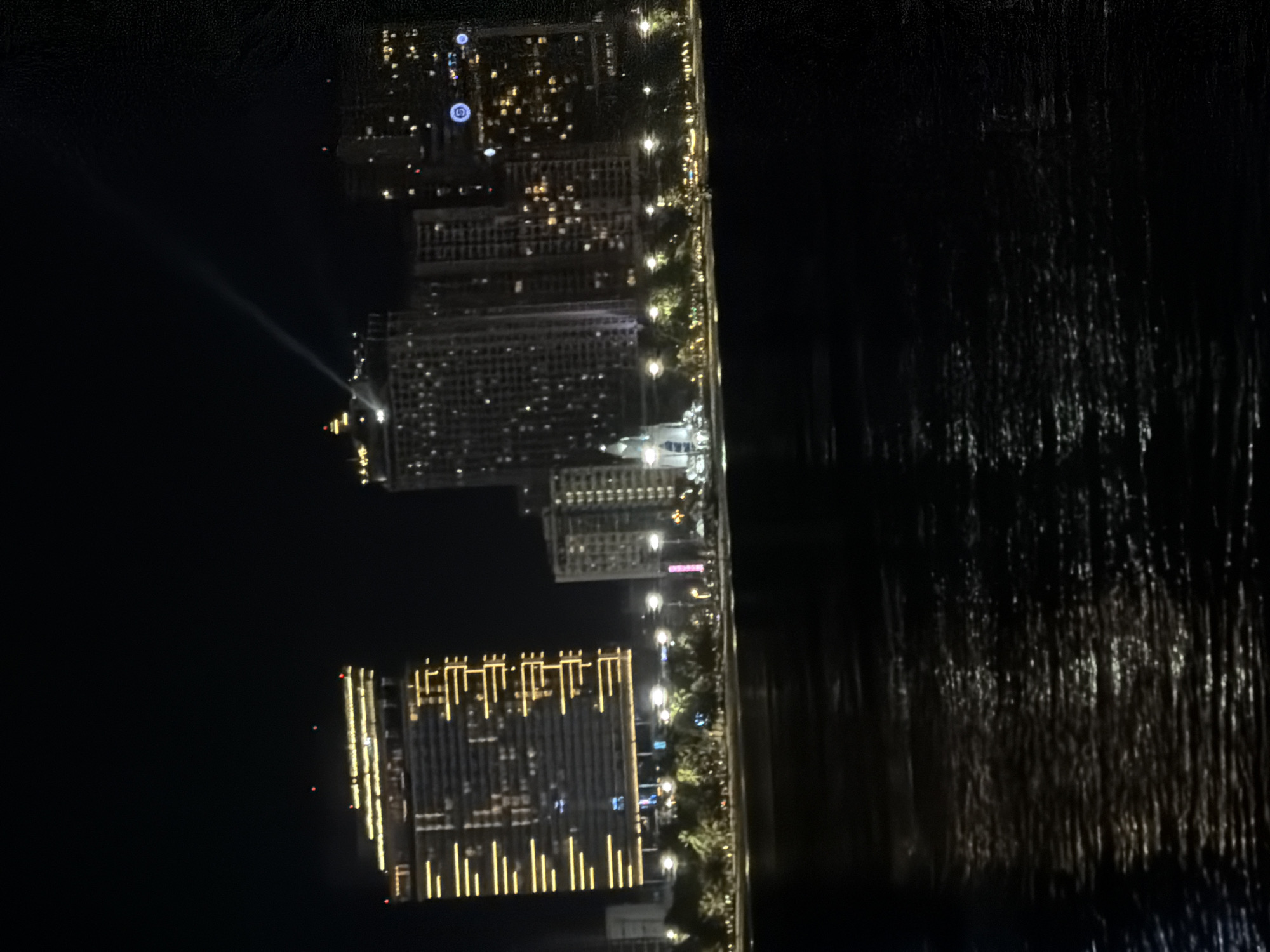 The image shows a nighttime cityscape in Nha Trang Ward, Vietnam, characterized by illuminated buildings and a waterfront. In the foreground, dark water reflects the city lights, creating a shimmering effect. Beyond the water, a coastline is lined with palm trees and streetlights. Several tall buildings are prominent, with the most striking being a large, modern skyscraper on the left, illuminated by horizontal yellow lights. To its right, a cluster of other high-rise buildings are visible, also lit up, suggesting a commercial or residential area. One building in the center has a distinct spire and appears to be a prominent landmark. A beam of light emanates from the top of one of the buildings, projecting upwards into the night sky. The overall scene depicts a vibrant urban environment at night, with the water serving as a reflective element connecting the city to the viewer. No people are discernible in the image.