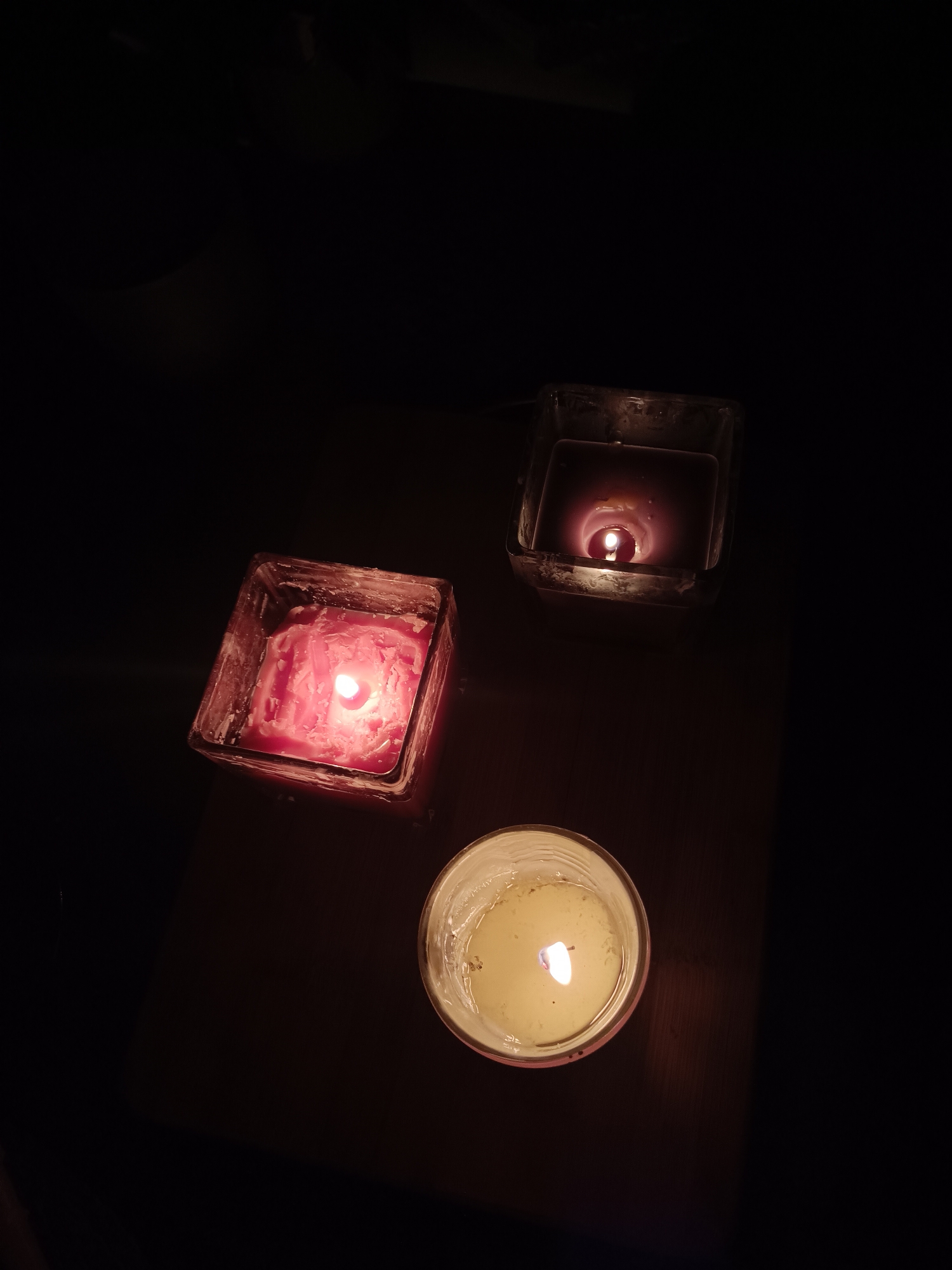 Three lit candles are arranged on a dark surface. The topmost candle is in a square glass holder and appears to be dark red or purple. The candle on the left is also in a square glass holder and is pink. The bottom candle is in a round glass holder and appears to be off-white or yellow. The surroundings are dark. The scene is not identifiable as being in Plovdiv, Bulgaria, or any specific location. There are no people or actions visible in the image.