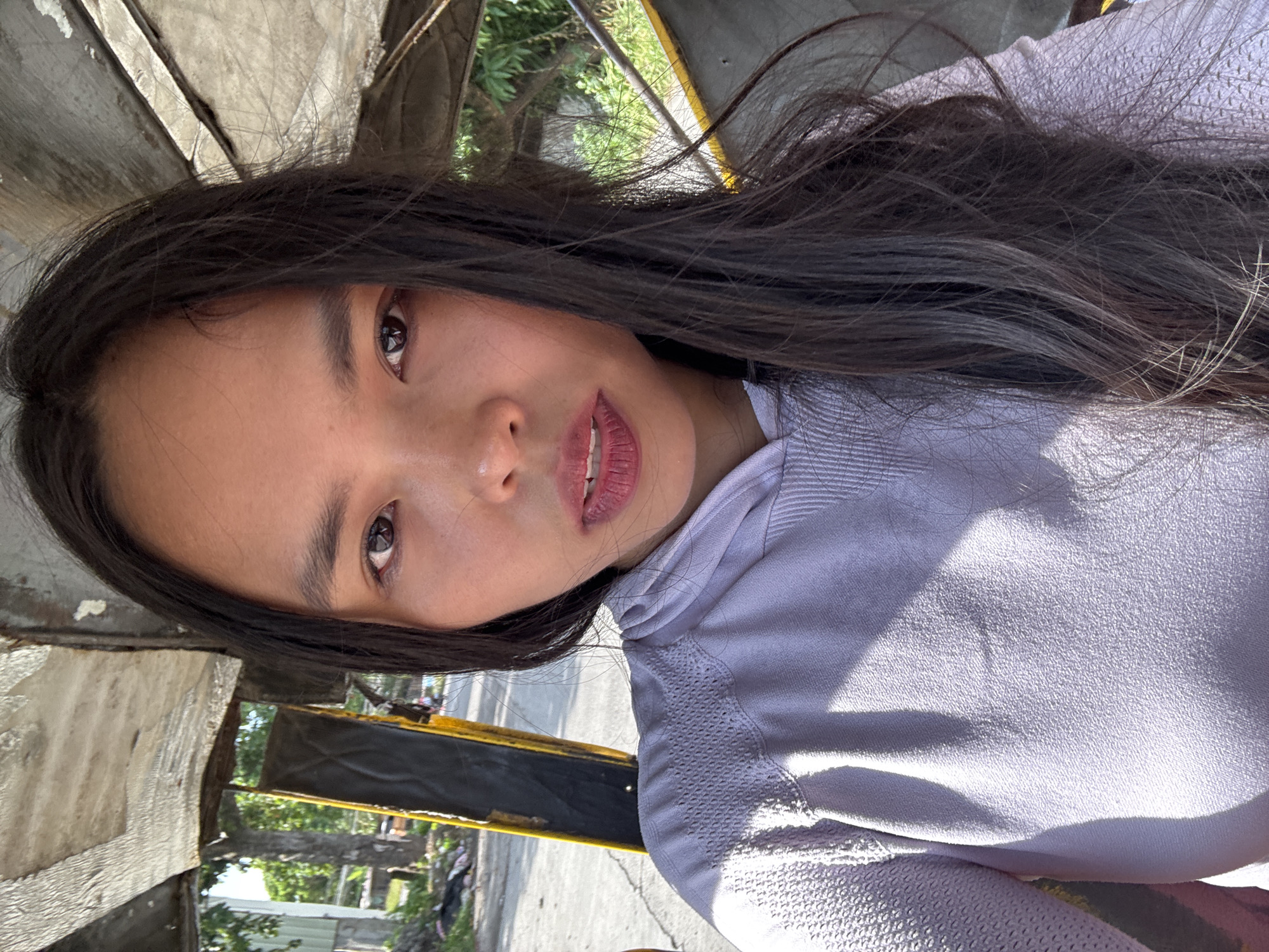 A young woman with long, dark hair is seen in a close-up selfie taken from inside a tricycle. She is wearing a light purple hooded sweatshirt with textured detailing on the shoulders and sleeves. Her face is centrally positioned in the frame, with her eyes looking directly at the camera. She has on lip gloss. The background shows the interior of a tricycle, including a portion of the roof and a yellow and black structure. Outside the tricycle, a blurred street scene with trees and buildings is visible. The image appears to be taken outdoors during the day, with natural sunlight illuminating the scene. The location is identified as Davao City, Philippines.