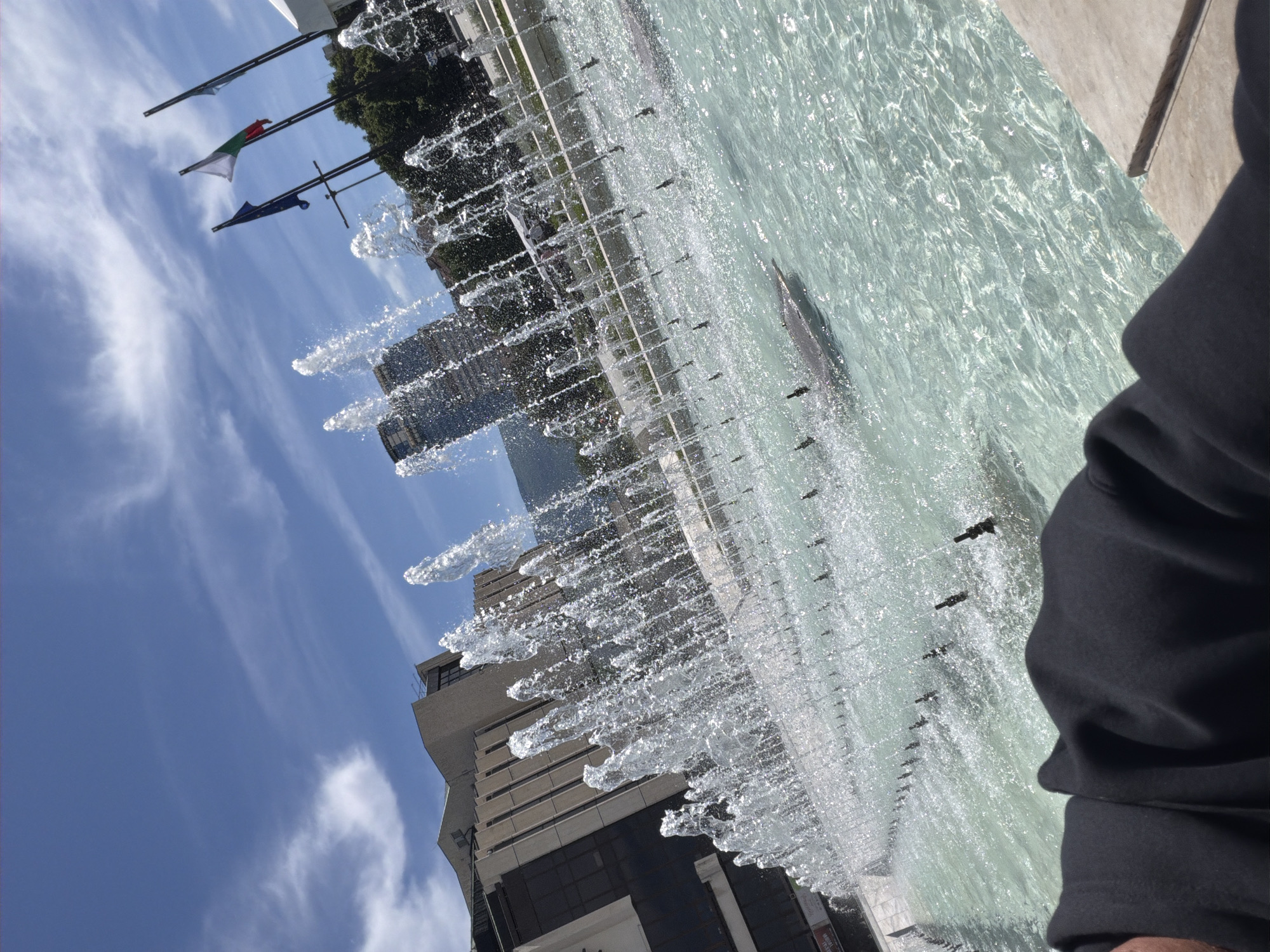 The image captures a cityscape scene featuring a fountain, flags, and buildings under a blue sky with wispy clouds. The fountain is active, with water shooting upwards and creating a shimmering effect. Several flags are visible atop poles, including one with a white, red, and green pattern, and another with a blue, white, and yellow design. Buildings of various heights and architectural styles are seen in the background, suggesting an urban setting. A portion of what appears to be a dark-colored garment or object is present in the foreground, partially obscuring the view of the fountain. The overall orientation of the image is slightly tilted.
