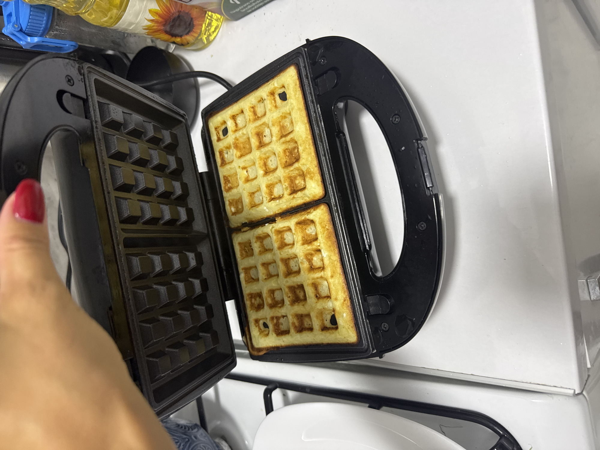 The image shows two golden-brown waffles being cooked in a black waffle iron. The waffle iron is open, and a hand with red nail polish is holding the lid. The waffle iron is sitting on a white surface, possibly a kitchen counter. In the background, there is a bottle of cooking oil with a sunflower on the label, and a blue water bottle. The scene appears to be in a domestic setting.