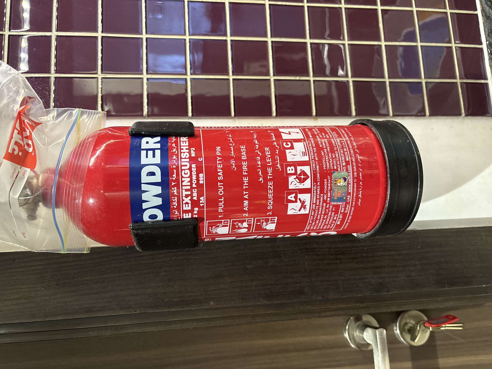 This media file shows a close-up, horizontal view of a red fire extinguisher. The extinguisher is branded "NAFFCO" and appears to be an ABC powder type, with a capacity of 2 kg. It is positioned on a white ledge, with a dark wooden surface in the foreground. In the background, a tiled wall with deep purple squares is visible.
The extinguisher has instructions printed on it in both English and Arabic, detailing how to use it: "PULL OUT SAFETY PIN," "AIM AT THE FIRE BASE," and "SQUEEZE THE LEVER." There are also symbols indicating its suitability for A, B, and C class fires. Visible text includes "POWDER," "EXTINGUISHER," "2 kg," "ABC POWDER," "MANUFACTURING FZCO," "Dubai UAE," and "MADE IN UAE." A plastic bag is partially visible on the left, covering part of the extinguisher.
The scene appears to be indoors, likely in a domestic or commercial setting given the presence of the tiled wall and wooden surface. The lighting suggests it is daytime. The overall impression is one of preparedness and safety equipment.