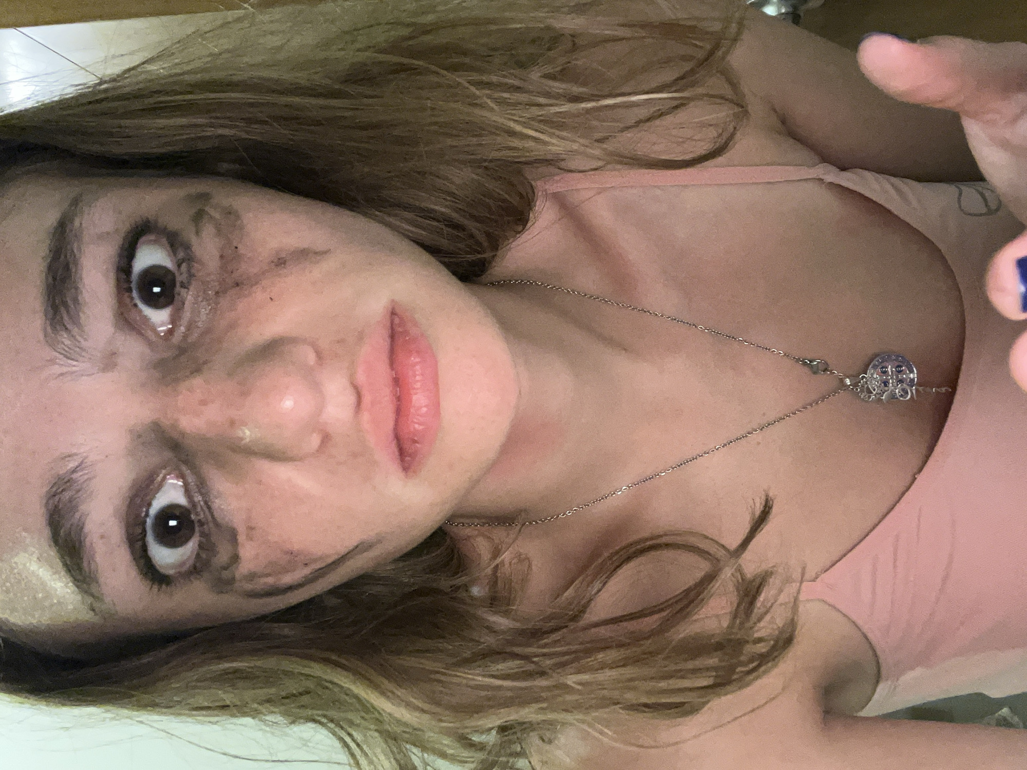 A woman with disheveled blonde hair is facing the camera with a concerned expression. There are streaks of dark smudges around her eyes and on her face, suggesting smeared makeup or dirt. She wears a pink tank top and a silver necklace with a pendant. Her left hand is partially visible in the upper right corner, with blue nail polish. The background is blurred. Location: San Bernardino Tlaxcalancingo, Mexico is not discernible in the image.