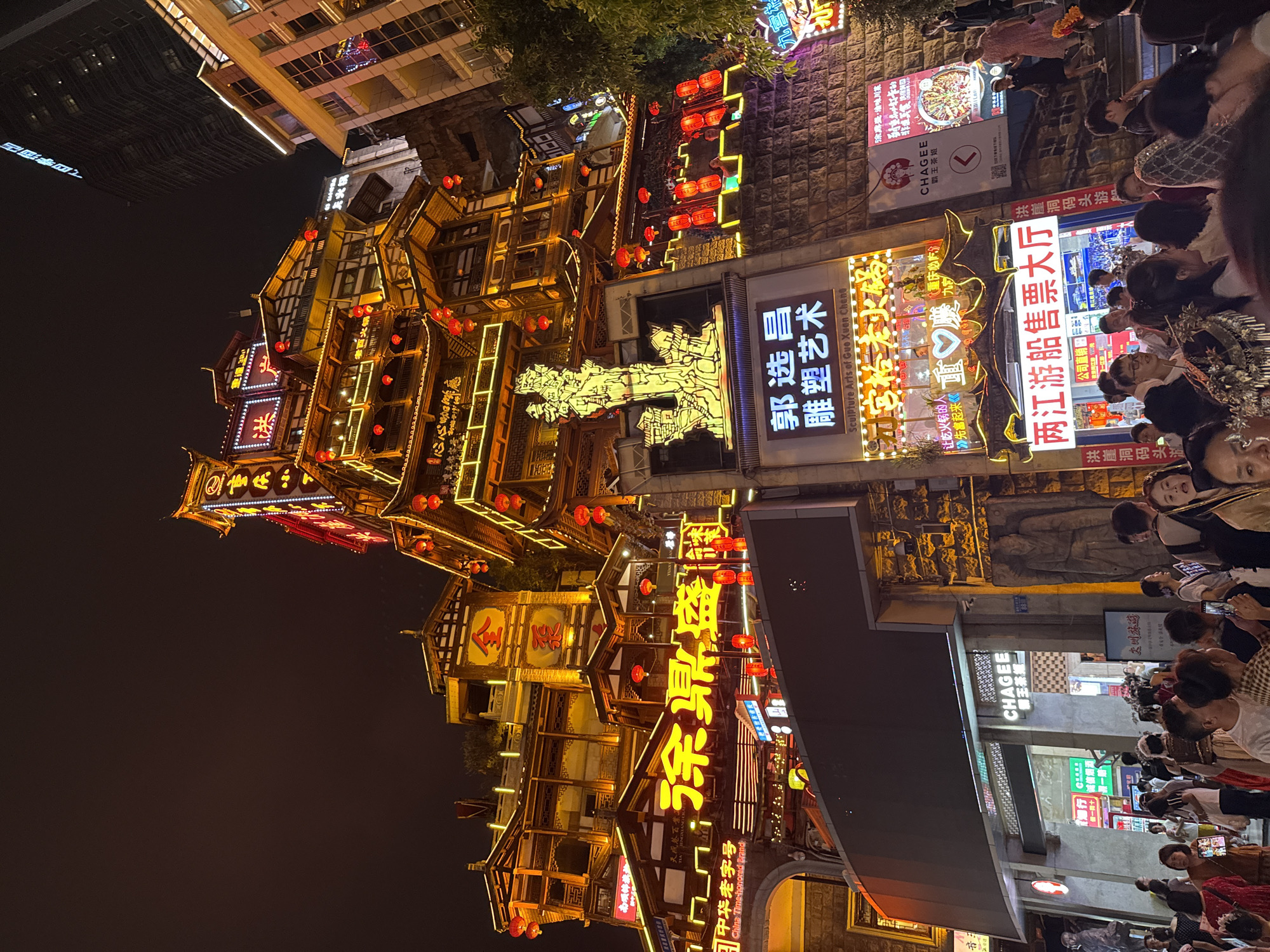 This is a nighttime exterior shot of a vibrant, multi-story building complex adorned with traditional Chinese architecture and illuminated by numerous neon signs and red lanterns. The building's facade is a mix of wooden structures and stone walls, with many levels featuring balconies and intricate roof designs. Large, brightly lit Chinese characters are prominently displayed, advertising various establishments, including "Chagee" tea shop and "Sculpture Arts of Guo Xuan Cheng." Below the main building, a lively street scene unfolds with a crowd of people gathered. Many individuals are holding up their phones, suggesting they are taking pictures or videos of the illuminated structures and the bustling atmosphere. The overall impression is of a popular tourist destination or entertainment area in a Chinese city, likely a commercial district with a strong emphasis on traditional aesthetics. The location is identified as Yuzhong District, China.