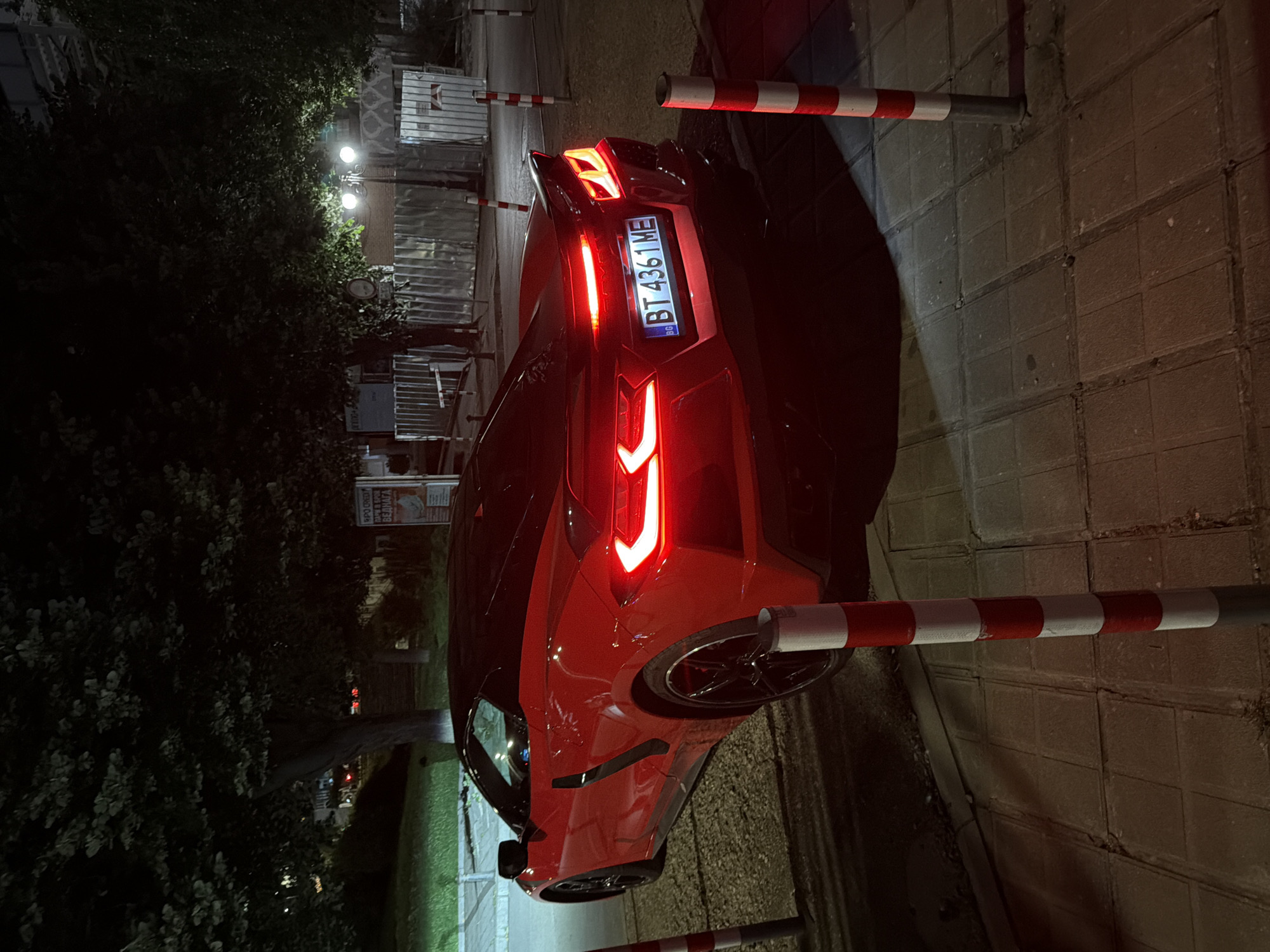 The image shows the rear view of a red Chevrolet Corvette C8 parked at night on a sidewalk in Veliko Tarnovo, Bulgaria. The car has its taillights illuminated. The license plate reads "BT 4361 ME" and has the Bulgarian national identifier "BG" on the left side. The sidewalk is paved with rectangular tiles. Red and white striped bollards are visible in the foreground and along the side of the car. Behind the car, there are trees with dense foliage and streetlights casting a warm glow. In the background, there are buildings and temporary construction barriers made of corrugated metal sheets. A sign with Cyrillic text, likely an advertisement, is partially visible on the right side of the image. The overall scene suggests a street in a city at night, with the car being the primary subject of interest.