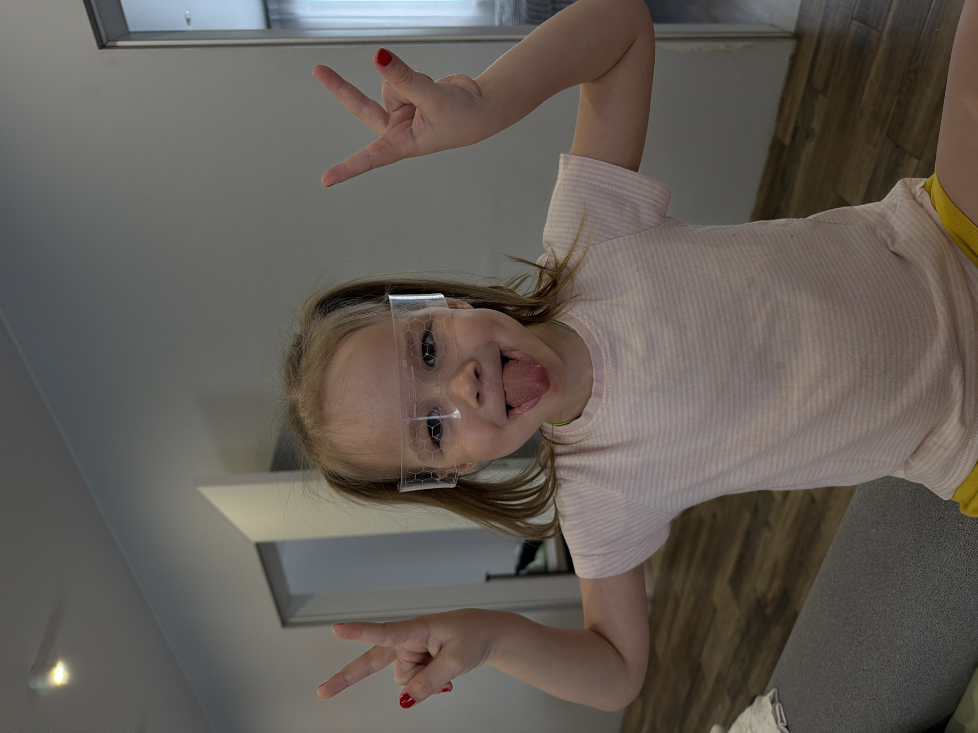 A young child with blonde hair, wearing a striped pink shirt, yellow shorts, and clear plastic glasses with a hexagonal pattern, is making a silly face with her tongue sticking out. She is giving a "peace" sign with both hands, and her fingernails are painted red. The background includes a ceiling with a recessed light fixture, a door, a section of wooden flooring, and a gray sofa. It is not possible to verify from the image alone that the location is Chaikovsky, Russia.