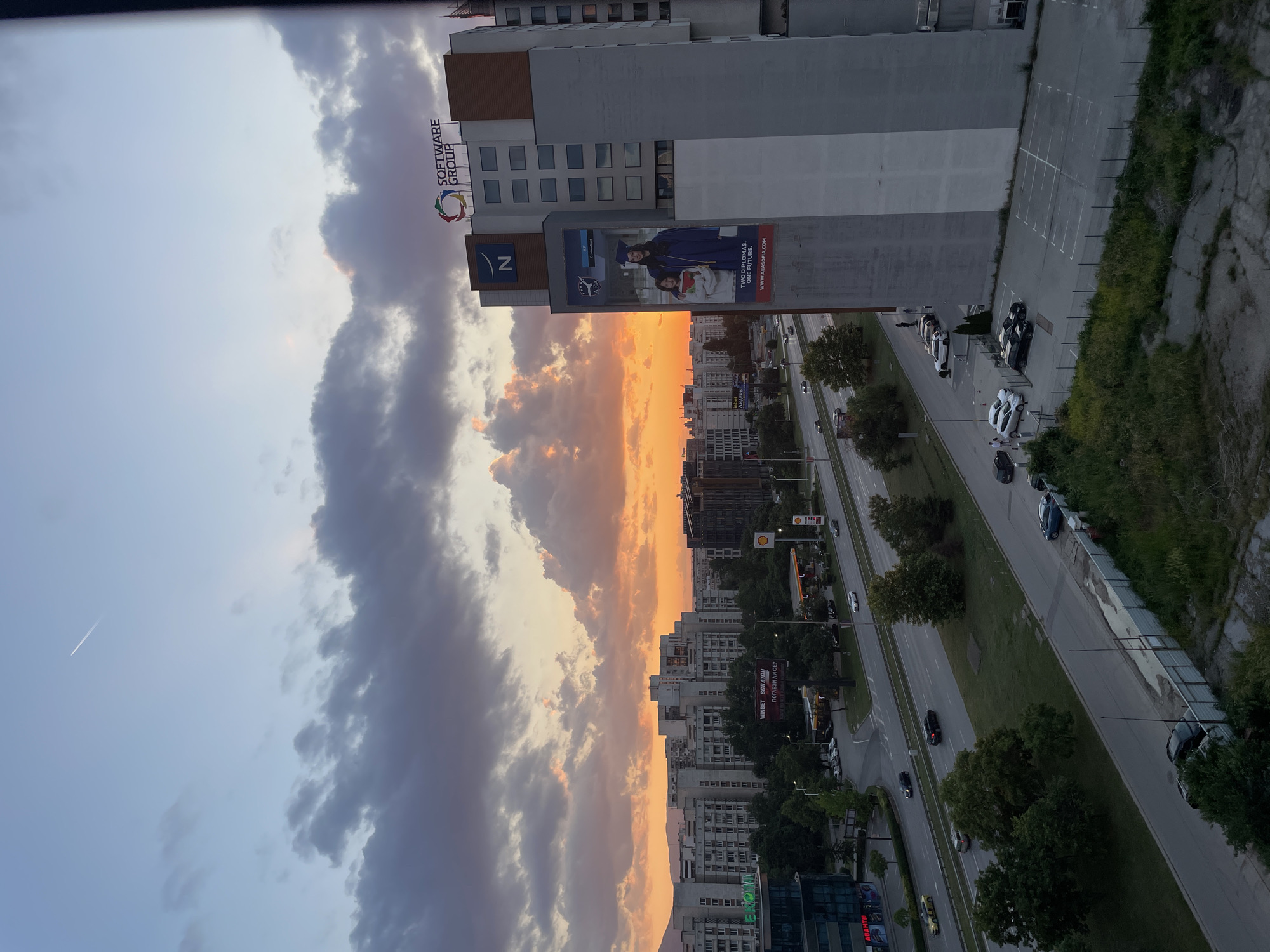 This image presents an urban landscape in Sofia, Bulgaria, at sunset, viewed from an elevated position. The sky displays a gradient of colors, transitioning from light blue at the zenith to vibrant orange and red hues towards the horizon, indicating the setting sun. Dark, textured clouds are scattered across the mid-sky, partially illuminated from below by the sunset. A faint white contrail from an aircraft is visible in the upper-left quadrant of the sky.
In the midground, a multi-lane highway, identified as the Sofia Ring Road, traverses the scene horizontally, with vehicles traveling in both directions. Trees line segments of the highway and a central median. An intersection is visible to the left, leading to an area with parked cars and a Shell gas station. A large billboard near the gas station displays "WINBET SCRATCH" and Cyrillic text reading "ПОБЕЖДАЙ ЛИ СЕ?".
To the left of the highway, numerous multi-story residential buildings with uniform facades are prominent. Some commercial signs are visible on the ground level of these buildings, including "Ekont" and "АВАНТИ". Distant mountains are faintly discernible on the horizon beneath the sunset colors.
On the right side of the image, a tall, modern building with a multi-paneled facade of gray, white, and brown is a central feature. At its peak, a "SOFTWARE GROUP" sign with a colorful circular logo is affixed. Below this, a large blue "N" logo is present. A significant vertical banner on the building's side features a woman in a graduation cap and gown with a book, alongside a younger girl. Text on the banner identifies it as "AEA" (American English Academy Sofia), stating "TWO DIPLOMAS. ONE FUTURE." and "WWW.AEASOFIA.COM". This building is located within Business Park Sofia.
In the immediate foreground, a fenced-off grassy area with some sparse vegetation and concrete debris is visible. A paved parking lot with several parked cars and a few individuals is situated next to the base of the "SOFTWARE GROUP" building.
The scene depicts a typical evening rush hour or post-work period, with active traffic and an overall calm atmosphere under the dramatic sunset sky. The location is precisely identifiable as the area around Business Park Sofia, looking across the Sofia Ring Road towards the Mladost district.