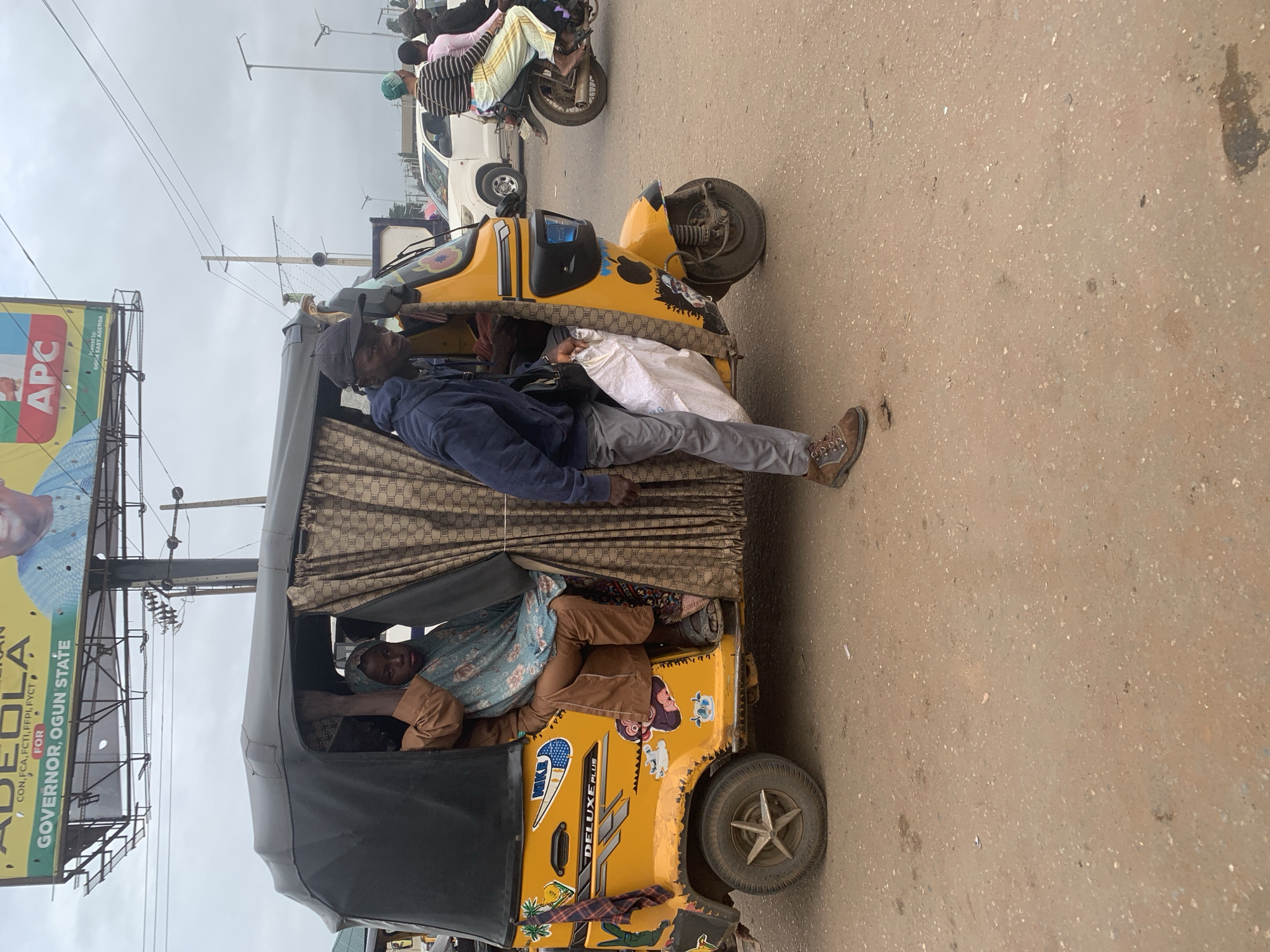 An eye-level, medium shot captures a yellow auto-rickshaw on a dirt road in Shagamu, Nigeria. The rickshaw, adorned with various stickers including "DELUXE PLUS" and a Nike logo, is partially covered by a patterned brown and gold curtain. Inside the rickshaw, a woman wearing a blue headscarf and brown attire sits to the left, while another person, whose face is obscured, is visible behind her. A man in a blue hoodie and grey pants stands beside the rickshaw, holding a white bag. In the background, a billboard for "ADEOLA" for "GOVERNOR, OGUN STATE" is visible, along with power lines and streetlights. Other vehicles, including a white car and a motorcycle with two riders, are present on the road. The sky is overcast.