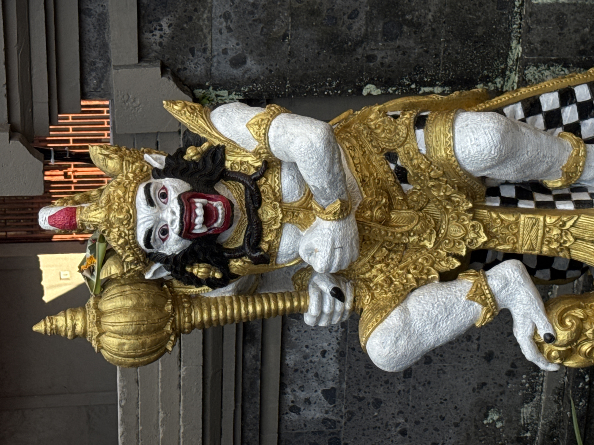A close-up, eye-level, vertical outdoor shot features a statue of a mythical creature, likely a guardian figure, with white skin and ornate gold armor and headdress. The creature has a fierce expression with wide, red-rimmed eyes, black hair, and an open mouth with sharp teeth, revealing a red interior. It wields a large, golden club-like object in its left hand and its right arm is bent, with its fist clenched near its chest. The statue is positioned in front of a textured gray stone wall and a dark opening with a pattern of vertical orange bars, suggesting it is located at a temple or cultural site in Ubud District, Indonesia. The statue's lower half is adorned with a checkered black and white sarong-like garment.
