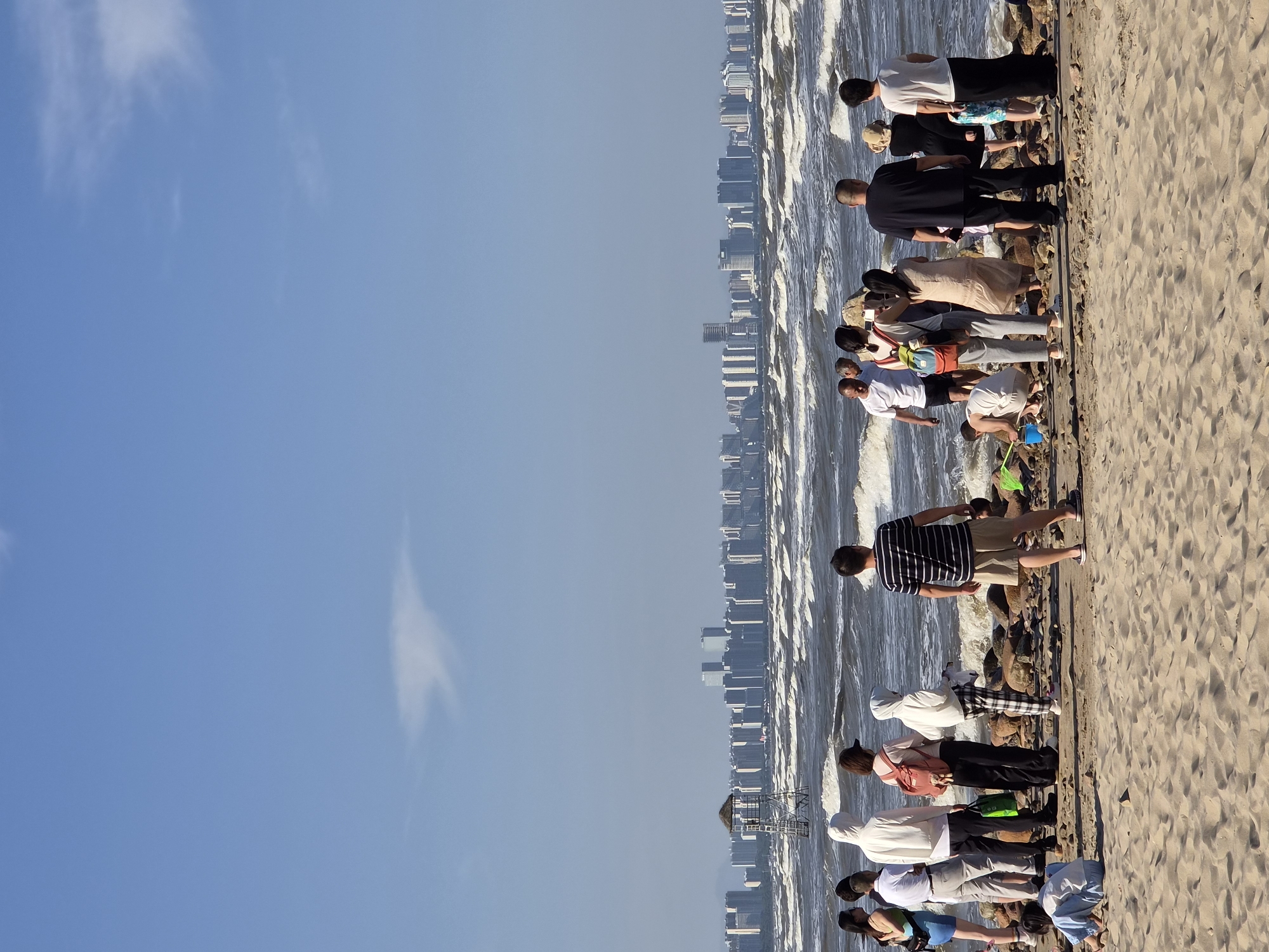 A group of people stand on a sandy beach in Qinhuangdao, China, looking out at a rough sea with a city skyline in the distance. The water is choppy, with visible waves breaking on a rocky shore. Several people are gathered in small groups, some appearing to be engaged in conversation, while others are looking at their phones or observing the water. One person is crouched down on the beach, seemingly playing in the sand. A lifeguard tower with a thatched roof stands on a rocky outcrop in the water. The sky is a clear blue with a few wispy clouds. The city skyline is composed of numerous tall buildings, indicating an urban area adjacent to the coast. The overall impression is of a recreational scene on a breezy day at the beach.