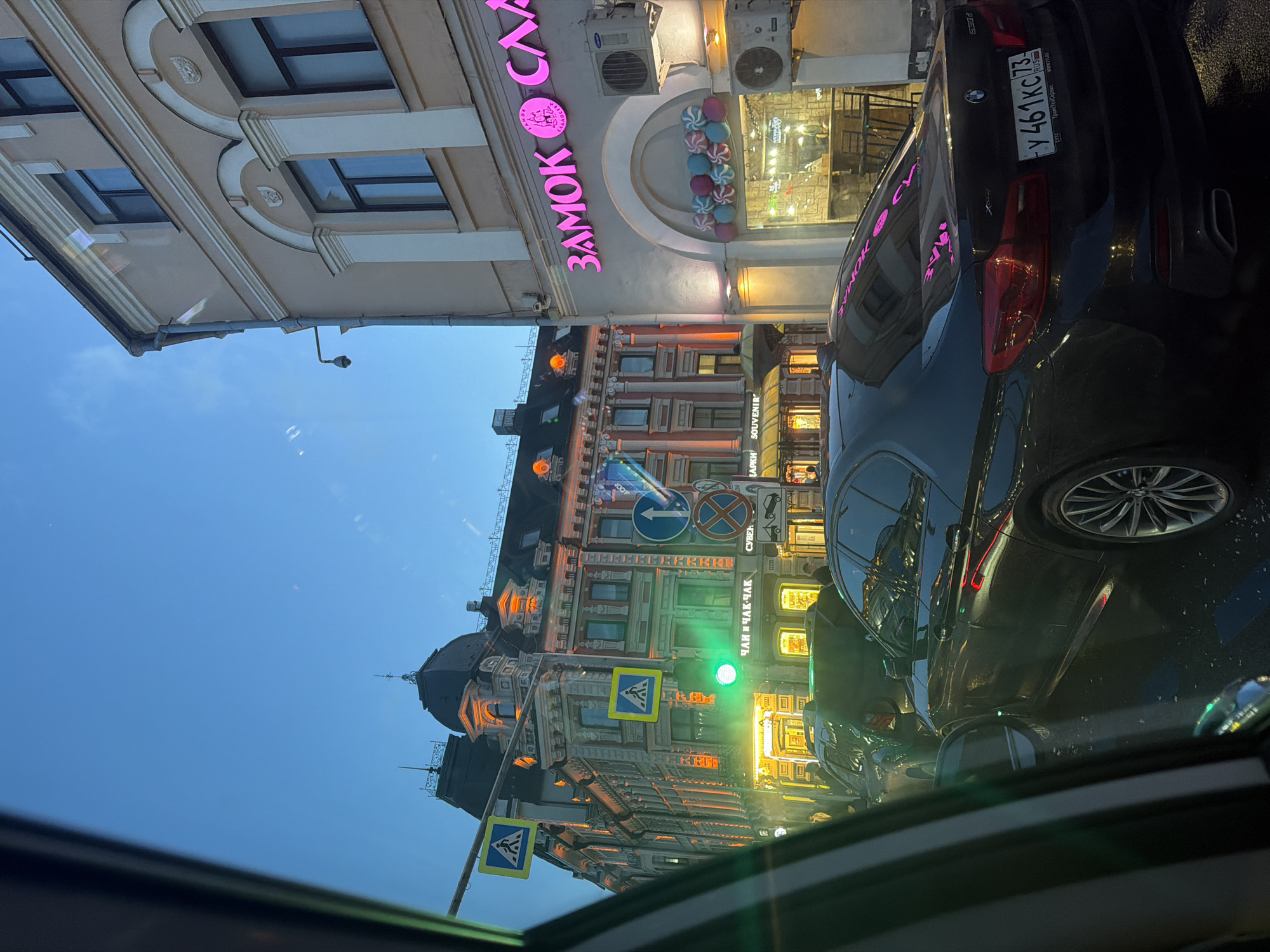 The image captures a vibrant street scene in Kazan, Russia, at dusk or early night, viewed from inside a car. The street surface appears wet, possibly from recent rain. A black BMW X3 SUV occupies the right foreground, stopped in traffic, with its rear license plate "у 461 КС 73 RUS" clearly visible. The plate also shows "ТрансТехСервис" and "WWW.TTS.RU". Its glossy surface reflects the illuminated surroundings.
On the left, a cream-colored building features ornate architectural details and a prominent pink neon sign that reads "ЗАМОК СЛА" (ZAMOK SLA), next to a display of colorful, spherical candies. Two air conditioning units are mounted above the sign. Further down the street, a more elaborate, older-style building with multiple windows, balconies, and possibly domes is brightly lit, showcasing signs that read "ЧАЙ И ЧАК-ЧАК СУВЕН" (CHAY I CHAK-CHAK SUVEN - Tea and Chak-chak Souvenirs) and "LAPKA SOUVENIR".
Several street signs are visible, including multiple yellow pedestrian crossing signs, a blue arrow indicating a left turn, and a red circular prohibitory sign. A green traffic light is also visible in the mid-ground, suggesting the vehicles are currently stopped at an intersection. The scene conveys the bustling atmosphere of a city evening, with commercial establishments glowing and traffic moving slowly. No specific individuals are discernible.