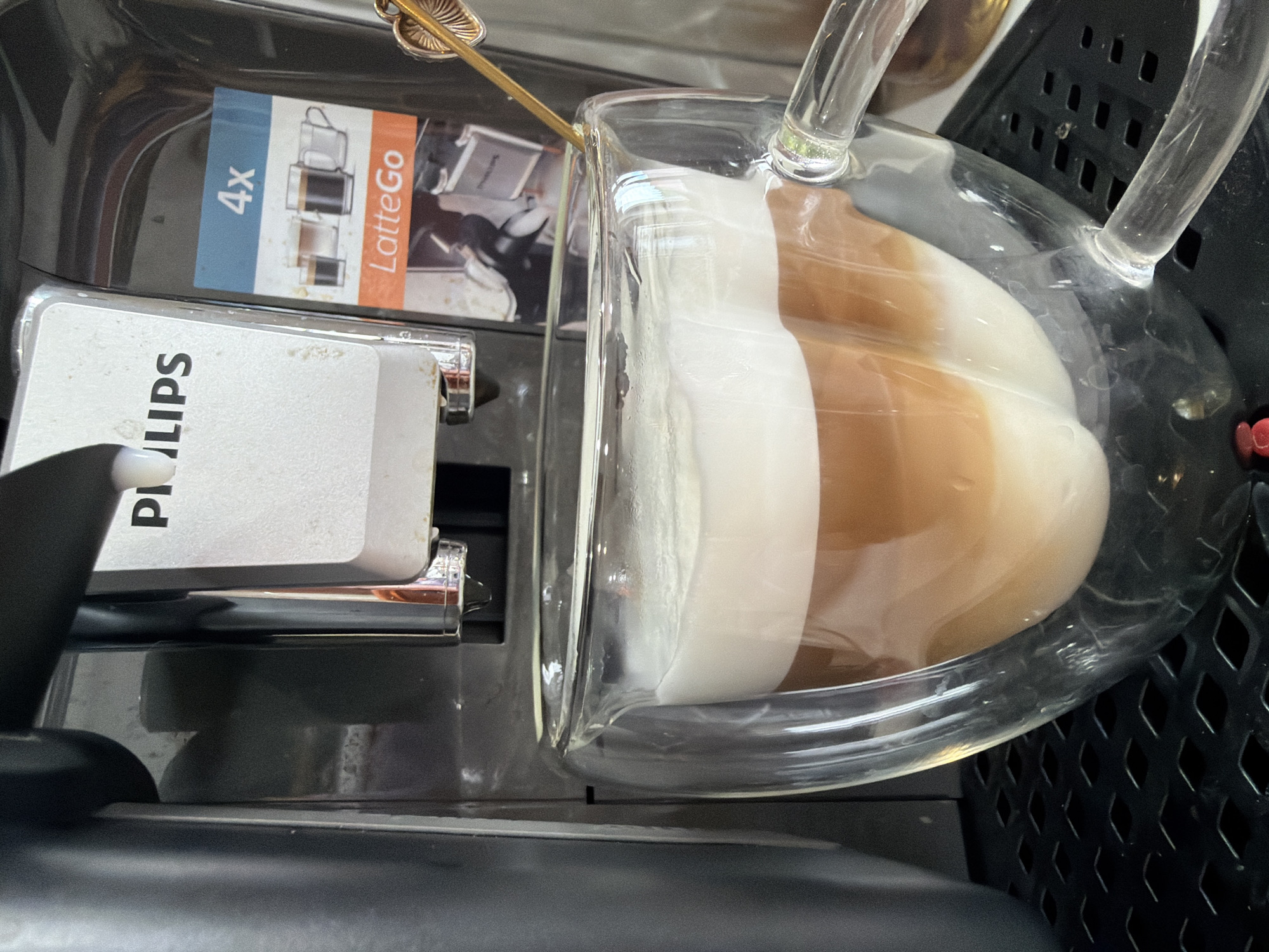 A close-up, high-angle shot captures a transparent, double-walled glass mug filled with a layered coffee drink, positioned on the drip tray of a Philips coffee machine featuring the "LatteGo" system. The coffee machine, predominantly dark grey with a prominent silver-colored dispensing unit bearing the "PHILIPS" logo, is actively dispensing or has just finished dispensing, as evidenced by a water droplet at the tip of the chrome spout. A promotional sticker on the machine indicates "4x LatteGo" and displays images of various coffee types.
The glass mug contains three distinct layers: a bottom layer of light beige liquid, a middle layer of darker brown coffee, and a substantial top layer of white milk foam. A golden-colored spoon with an ornate, leaf-shaped handle is partially submerged in the foam, leaning against the side of the mug. The mug rests on a black, textured drip tray characterized by a pattern of small, diamond-shaped perforations. The immediate environment appears to be an indoor setting, likely a kitchen or office space. No individuals are present in the image. The scene is located in the city of Chaikovsky, Russia.