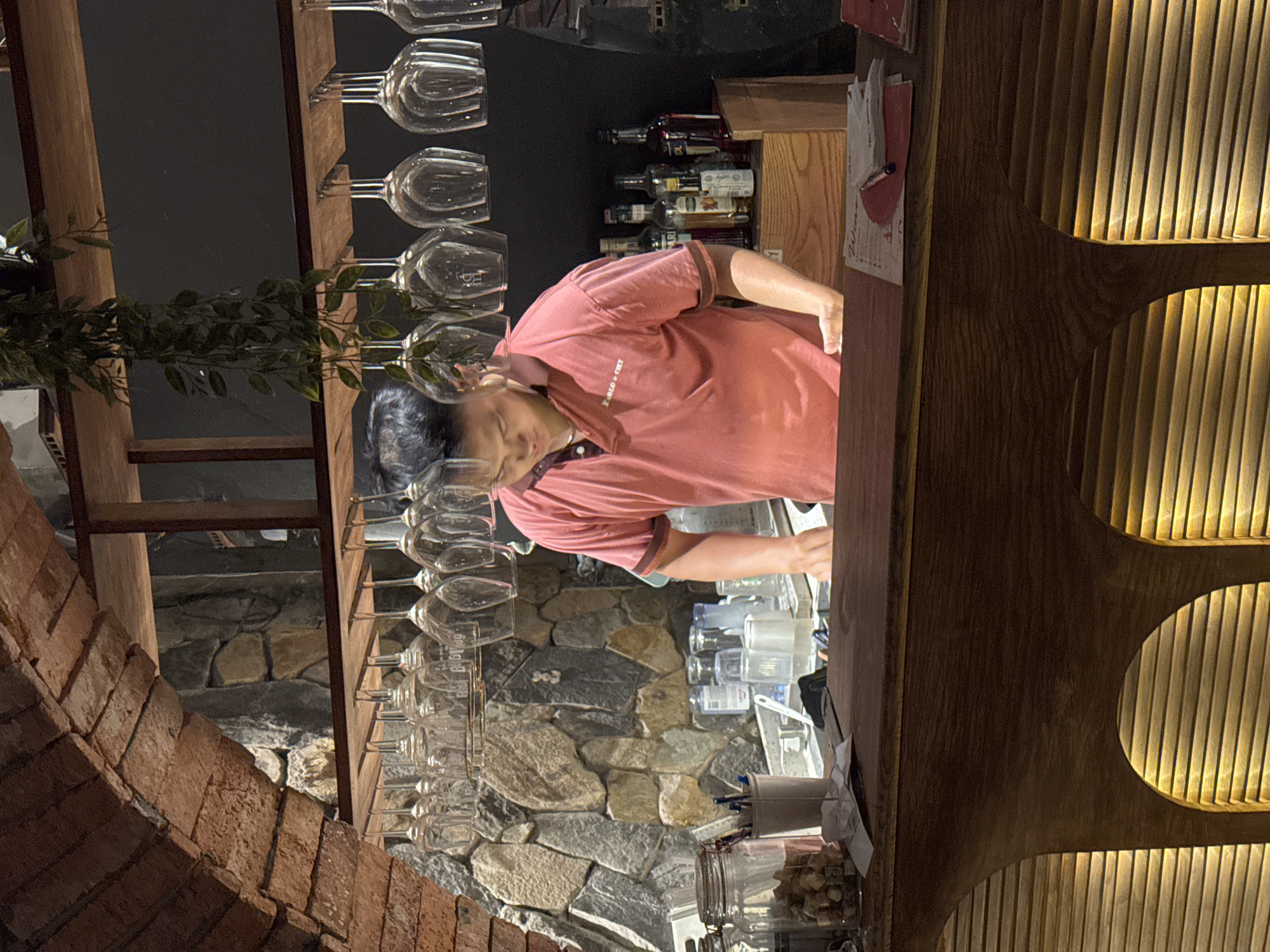 A male bartender, wearing a coral-colored polo shirt with a collar and ribbed cuffs, is pictured behind a wooden bar counter. He is leaning forward, appearing to be occupied with tasks related to his work. The bar is equipped with rows of inverted wine glasses hanging from a shelf above. Behind the bartender, a wall is covered with various sizes of rough-cut stones, creating a rustic backdrop. The bar area is illuminated by warm, ambient lighting, casting a soft glow on the wooden surfaces and the stone wall. The overall scene suggests an indoor bar or restaurant setting, possibly with a focus on wine service given the abundance of glassware. The image does not provide specific visual cues to determine the exact location in Hanoi, Vietnam, but the style of the bar and the decor could be indicative of contemporary establishments found in urban centers.