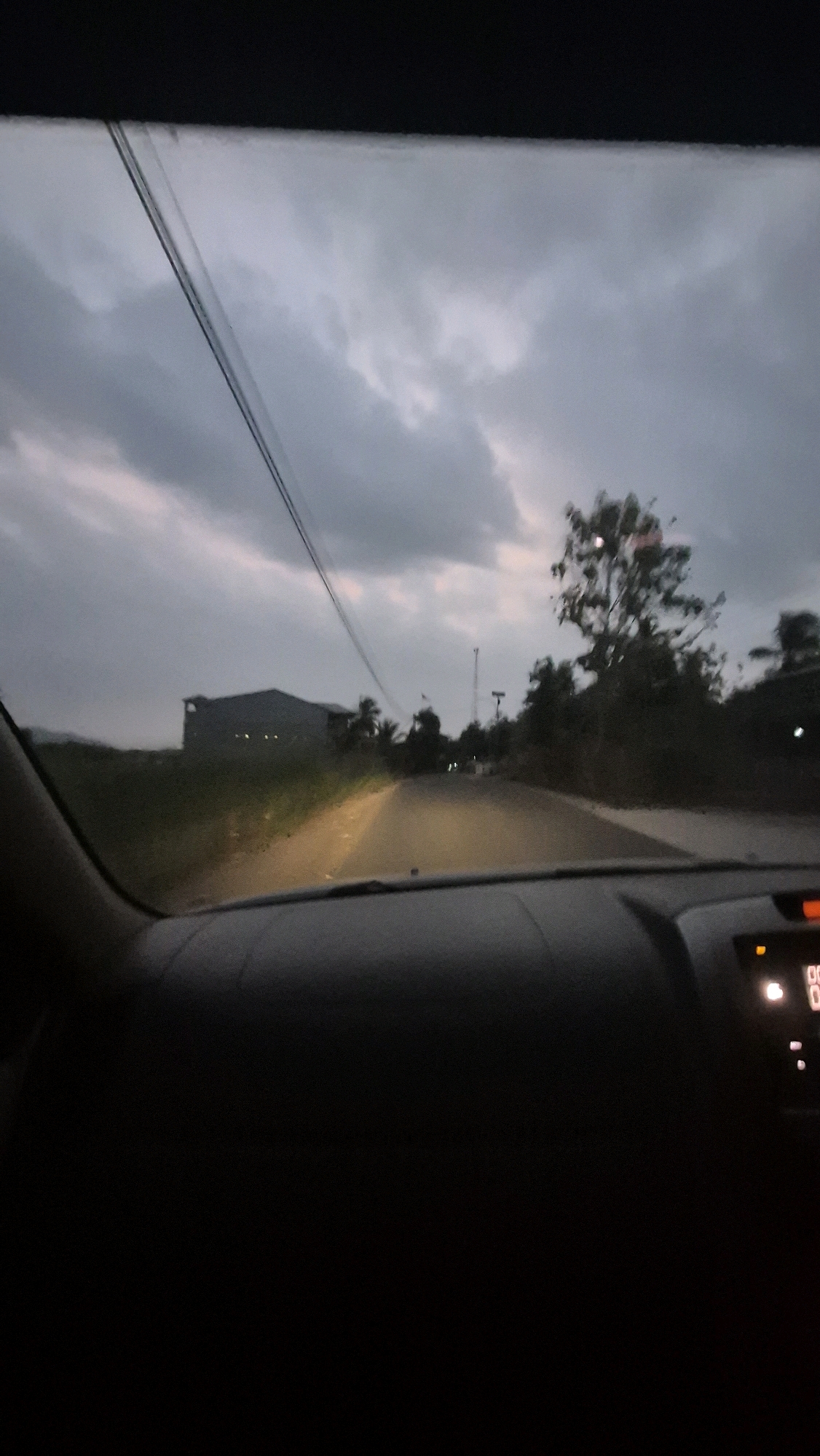 The image depicts a view from inside a vehicle at dusk, looking out at a road leading towards a large, dark building on the left and trees on the right. The sky is overcast with dramatic clouds. A power line runs diagonally across the upper portion of the frame. The road is illuminated by the vehicle's headlights. The interior of the car is dark, with a dashboard visible at the bottom. The scene appears to be set in a rural or semi-rural area, consistent with the location in Batuwarno, Indonesia, which is characterized by agricultural land and scattered settlements. There are no people or animals clearly visible in the image.