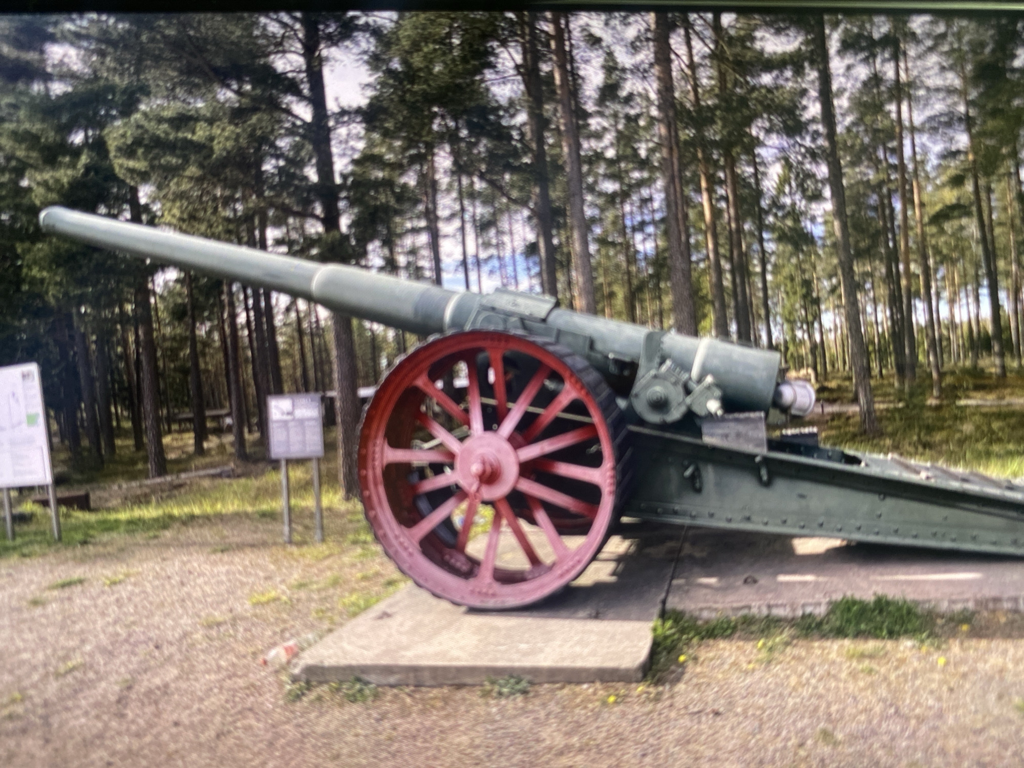 The image shows a large, green artillery piece positioned outdoors on a concrete slab, with a prominent red-spoked wheel. The artillery piece is angled diagonally, with its barrel extending upwards and towards the right. The background consists of a dense forest of tall, thin trees, with dappled sunlight filtering through the canopy. A gravel path with some sparse grass and yellow flowers is visible to the left of the concrete slab. A sign or informational board is partially visible in the upper right corner. This scene is located in Kerava, Finland.