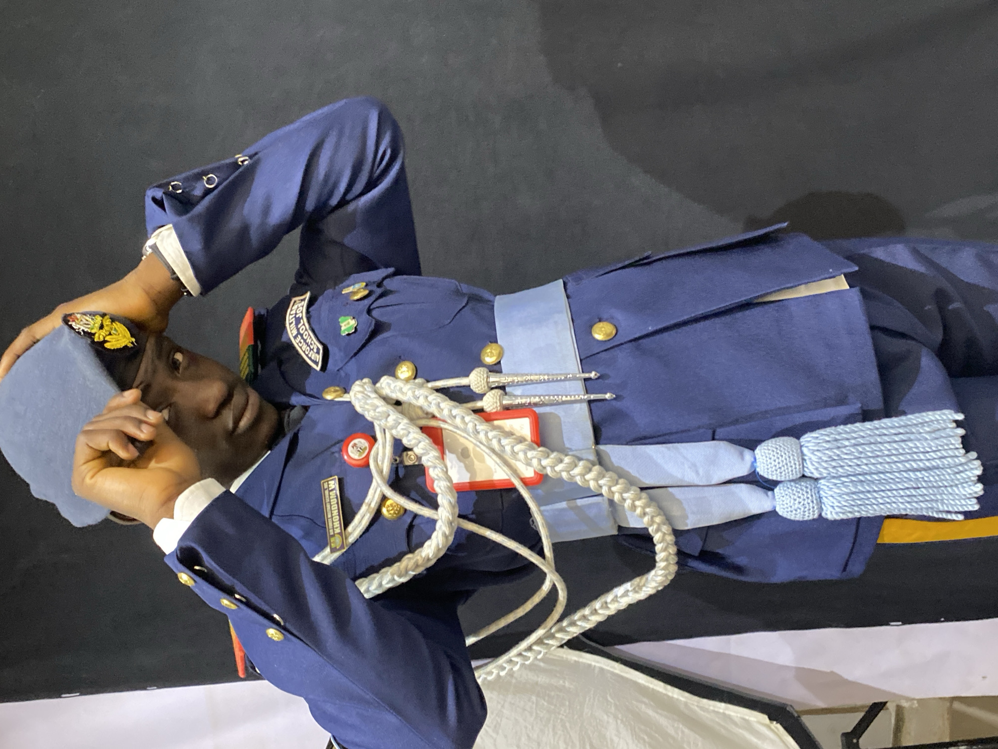 The image depicts a dark-skinned individual wearing a light blue military uniform. The uniform features gold buttons, badges, and a white rope cord with light blue tassels across the chest. The individual is also wearing a light blue cap with a gold emblem. A name tag on the uniform reads "KHONDUM W." In the background is a dark, indistinct area. Given the patch on the uniform referencing the "Air Force Military School Jos," the location is likely Jos, Nigeria. The individual is holding their head with their hand, as if in distress. The scene seems to be indoors.