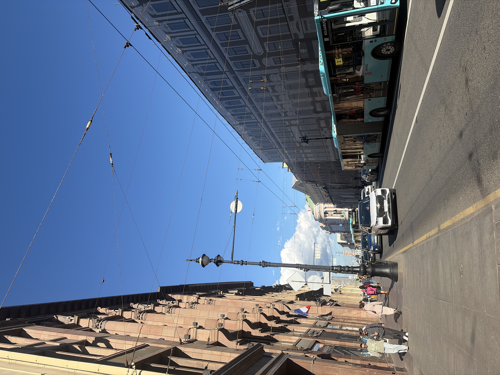 The image depicts a street scene in Saint Petersburg, Russia, on a clear, sunny day. The perspective is from a sidewalk, looking down a street lined with buildings. On the left, an ornate, multi-story building with classical architectural elements and decorative statues is visible. A French flag is mounted on this building. The sidewalk is made of stone paving and is populated by several pedestrians walking in both directions.
On the right side of the street, a modern, articulated blue bus is driving past. The bus has "VOLGABUS" branding. In front of the bus, a white SUV with a license plate that appears to be in Cyrillic script is also traveling on the road. Several other vehicles are visible further down the street. Overhead, numerous electrical wires for public transportation are suspended across the sky. The sky is a vibrant blue with a few scattered white clouds. The overall environment suggests an urban setting with active pedestrian and vehicular traffic.