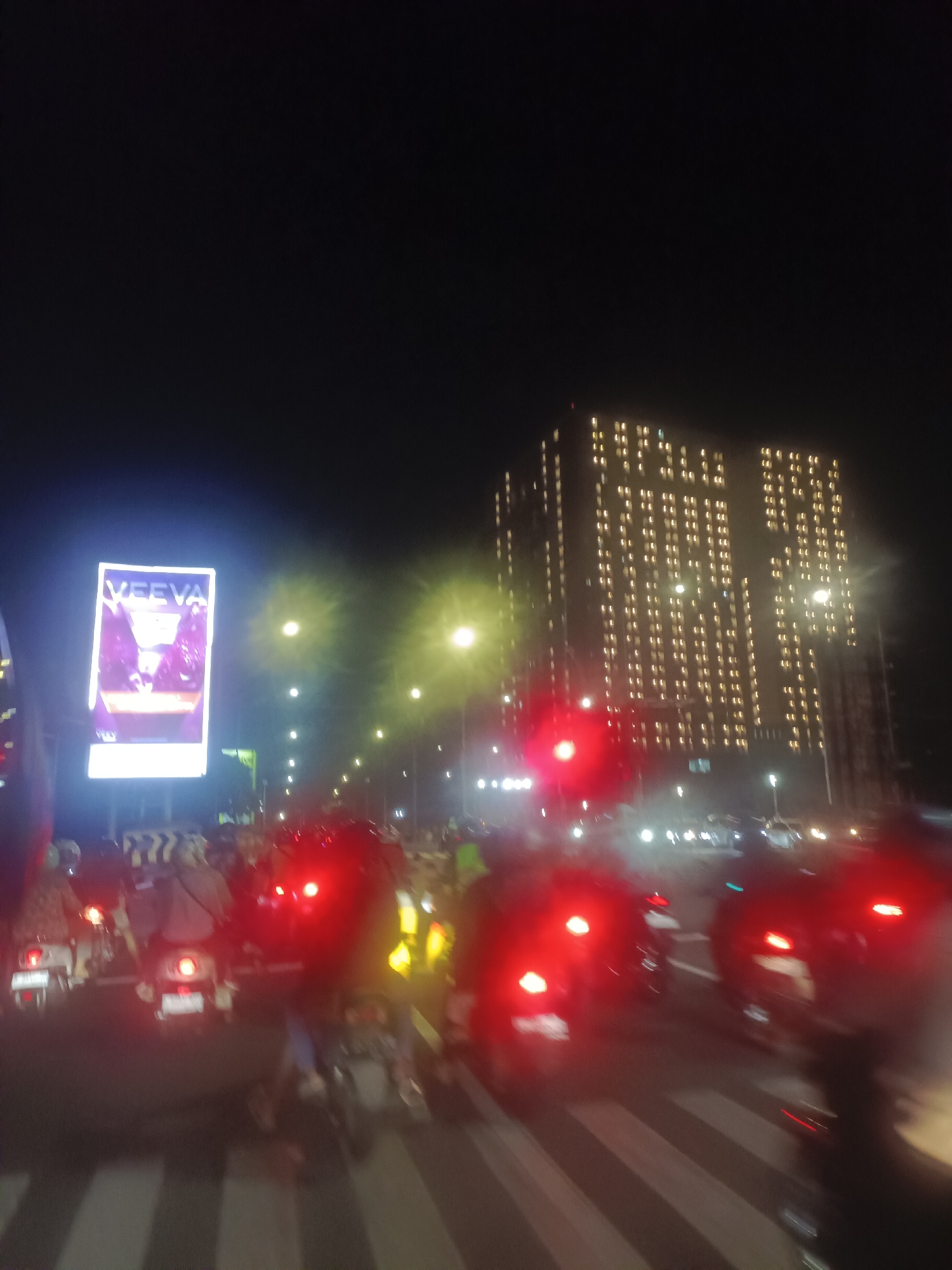 The image depicts a nighttime street scene in an urban environment, likely a city due to the presence of tall buildings and heavy traffic. The foreground is dominated by a row of motorcycles with illuminated red taillights, suggesting they are stopped in traffic, possibly at a zebra crossing visible beneath them. The image is somewhat blurry, indicating motion or a lack of focus.
In the background, a tall, modern building stands illuminated with numerous windows, suggesting it is either an apartment complex or an office building operating at night. To the left, a bright billboard displays the word "VEEVA" and an image, likely an advertisement. Streetlights are visible along the road, contributing to the overall illumination of the scene. The sky above is dark, indicating it is nighttime. The location is identified as the city of Unknown, Unknown.