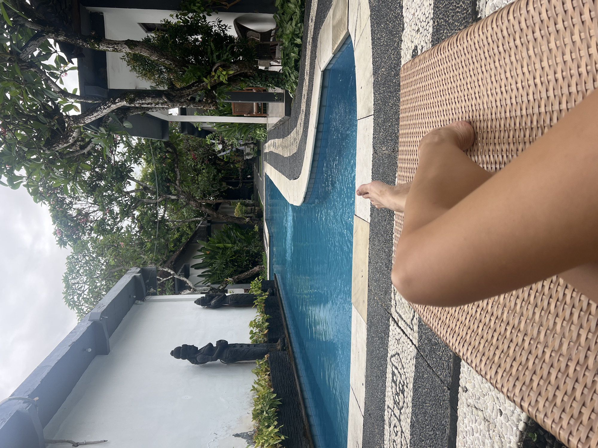 The image presents a first-person perspective of a tranquil poolside setting in Kuta Selatan, Indonesia. In the foreground, a person's legs and feet are visible, resting comfortably on a light brown, woven lounge chair. The chair is positioned on an intricately designed pebble mosaic floor featuring contrasting black and white stones that create decorative patterns around the swimming pool.
The scene features a clear, blue swimming pool, reflecting the overcast sky. Along the far edge of the pool, a row of dark, traditional Balinese-style stone statues stand on a low ledge, bordered by lush green foliage. Behind these statues, a white building wall with dark architectural elements is visible, partially obscured by tropical trees. The trees have thick, gnarled branches and abundant green leaves, characteristic of a tropical environment.
The sky is overcast, suggesting a cloudy day, possibly late morning or afternoon. The overall atmosphere conveys relaxation and an exotic resort or villa setting. No visible text is present in the image.