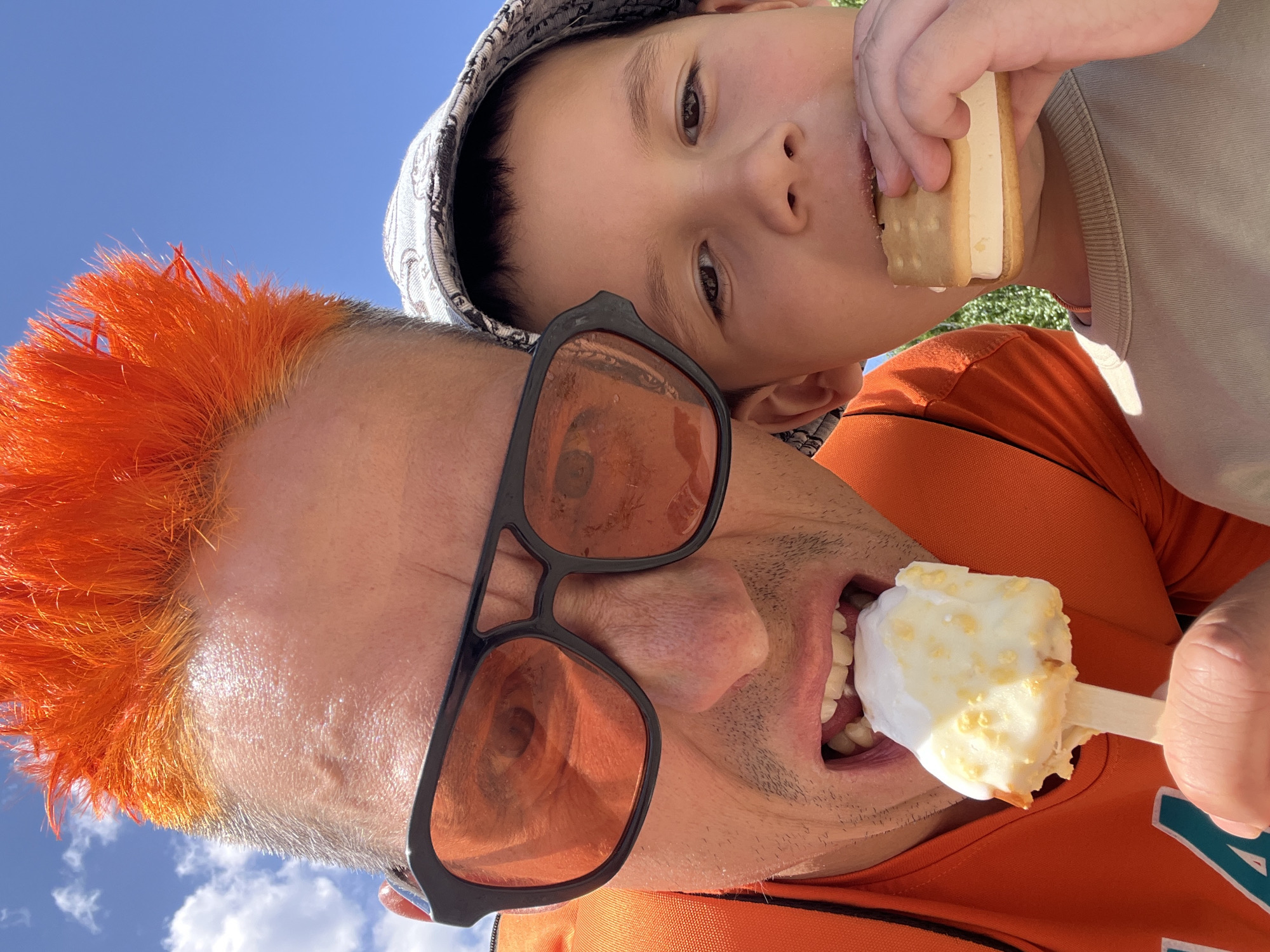 A selfie taken from a low angle shows a man and a young child outside under a blue sky with scattered white clouds. The man is in the foreground, wearing sunglasses with orange lenses and a black frame. He has short, orange-colored hair and is eating an ice cream popsicle on a stick. Behind him and slightly to the upper right, the child is wearing a patterned bucket hat and eating an ice cream sandwich. The man is wearing an orange shirt, and part of a light-colored shirt is visible on the child. The location is Chaikovsky, Russia.