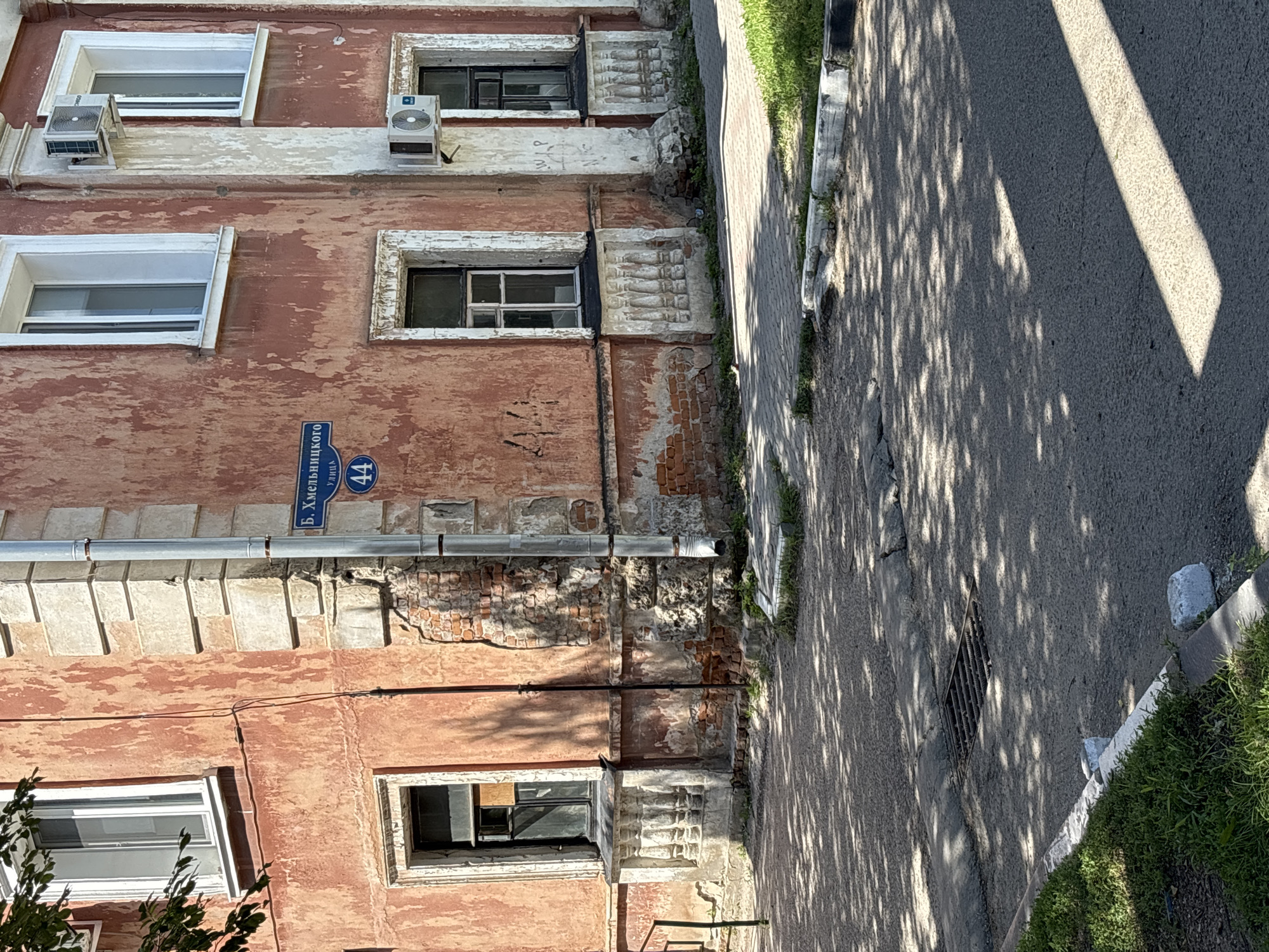 The image shows a faded reddish-orange building with chipped paint. The building has several windows, some with white frames and others with dark frames. There are also two air conditioning units mounted on the exterior wall. A street sign in Russian indicates "B. Khmelnytskogo Street 44." The building appears old and somewhat dilapidated. The foreground is a paved street with some grass and a storm drain. The scene is illuminated by sunlight, casting shadows from trees onto the street and building. There are no people or animals visible in the image. The location is identified as Blagoveshchensk, Russia.